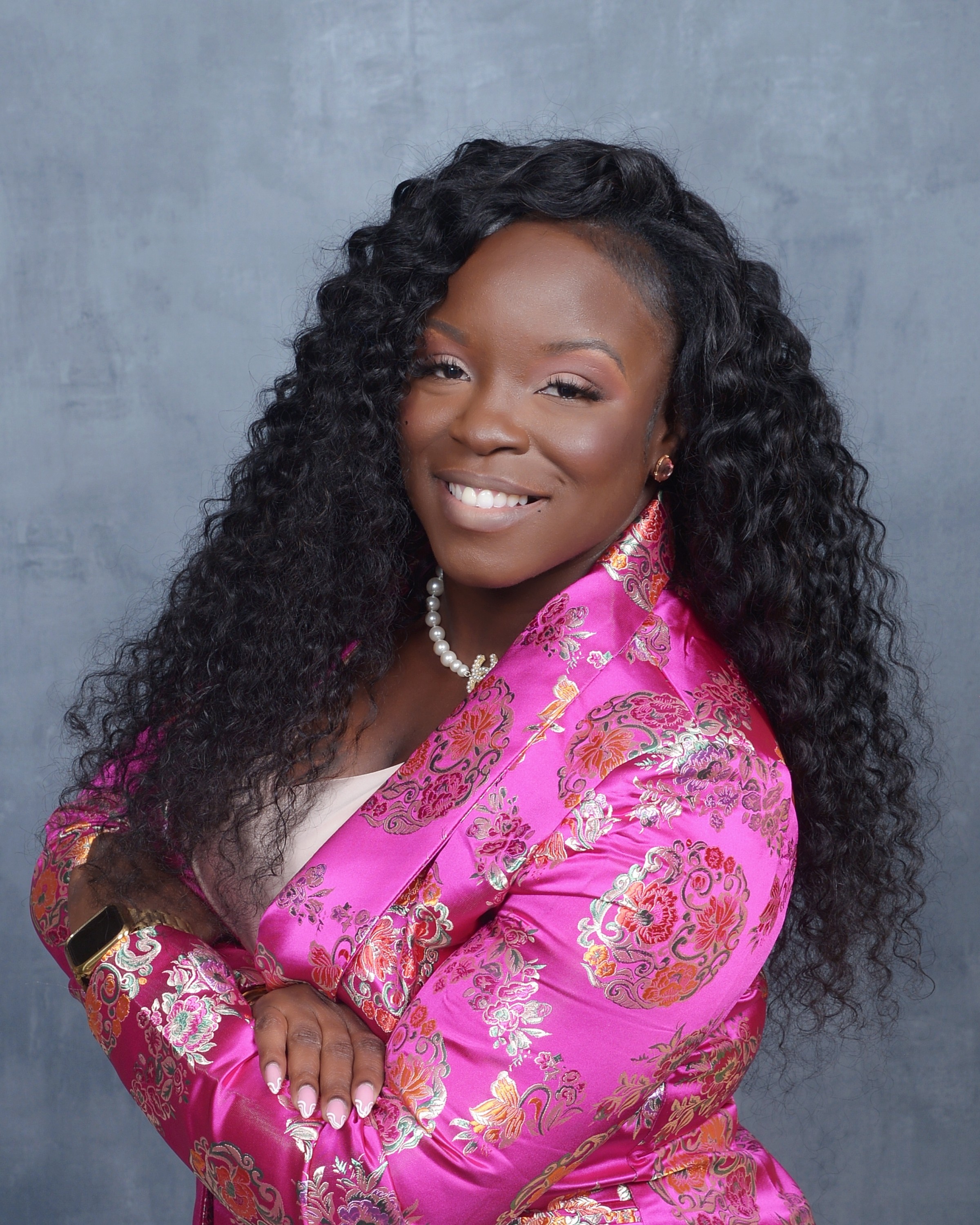 Woman with long curly hair smiling, wearing a pink and gold patterned jacket, pearl necklace, and earrings, against a gray background.