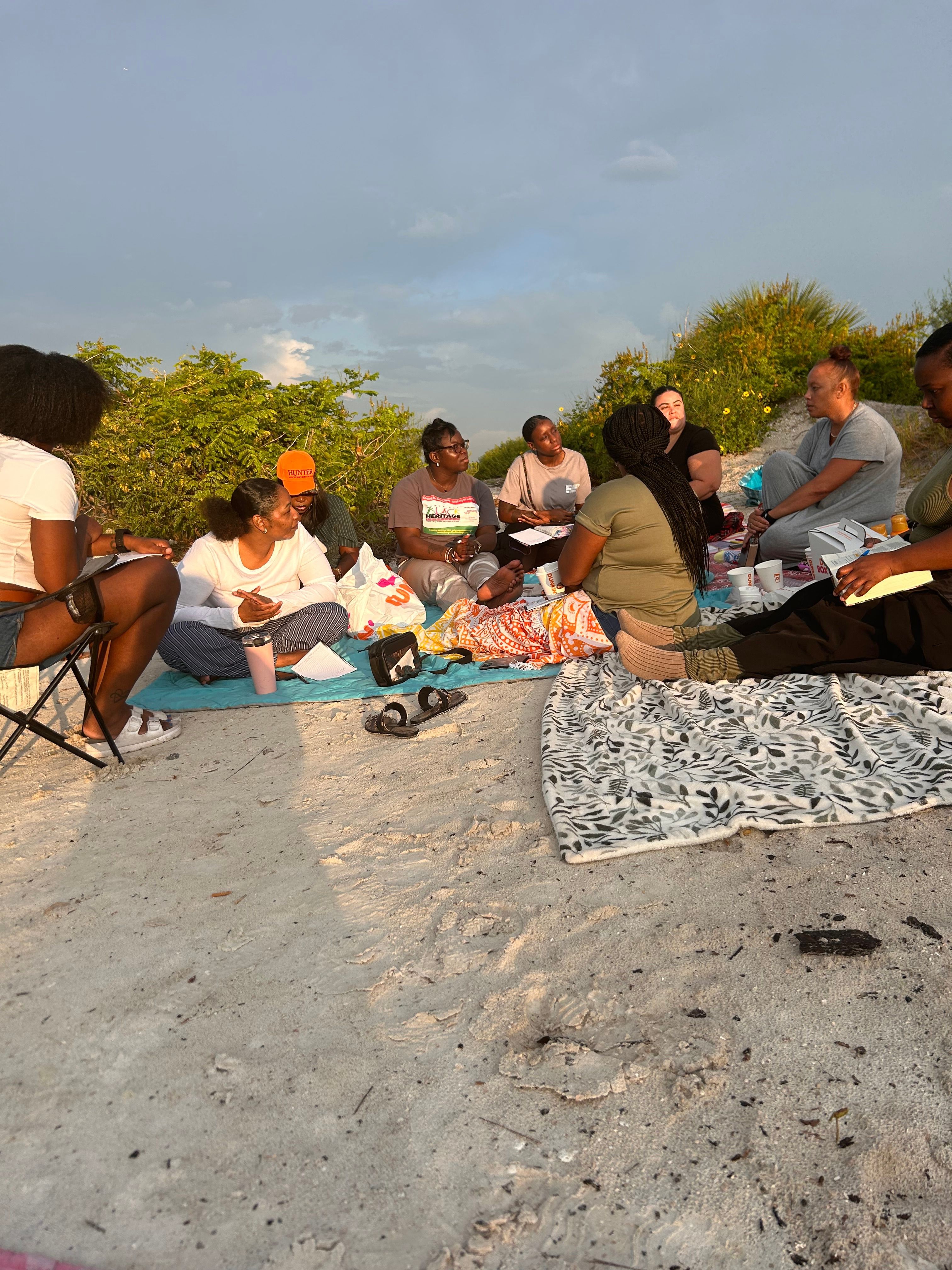 Group of people sitting on a beach, some with towels, with trees and sky in the background.