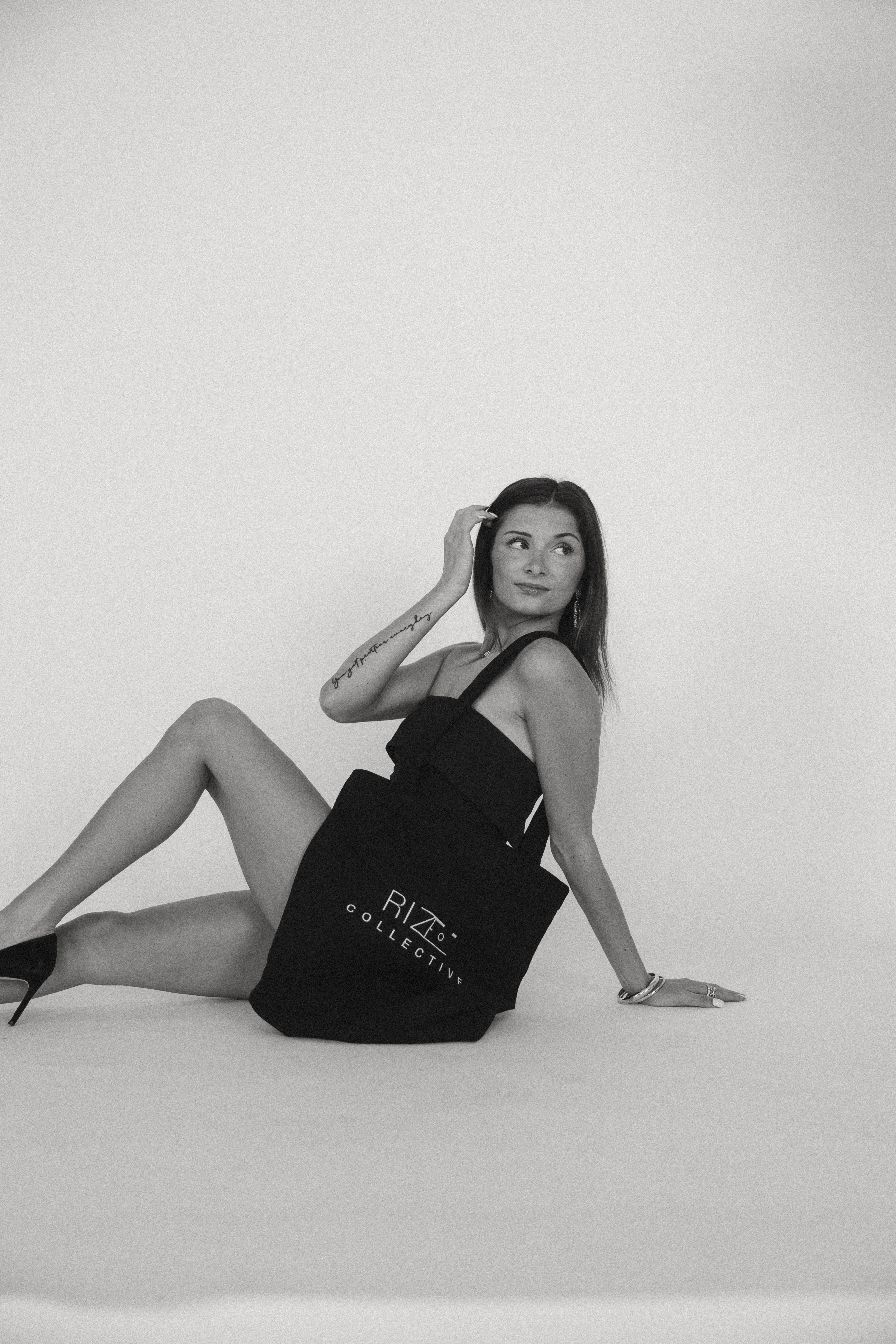 Woman in black dress sitting on the floor, posing with one hand on her head, against a plain background.