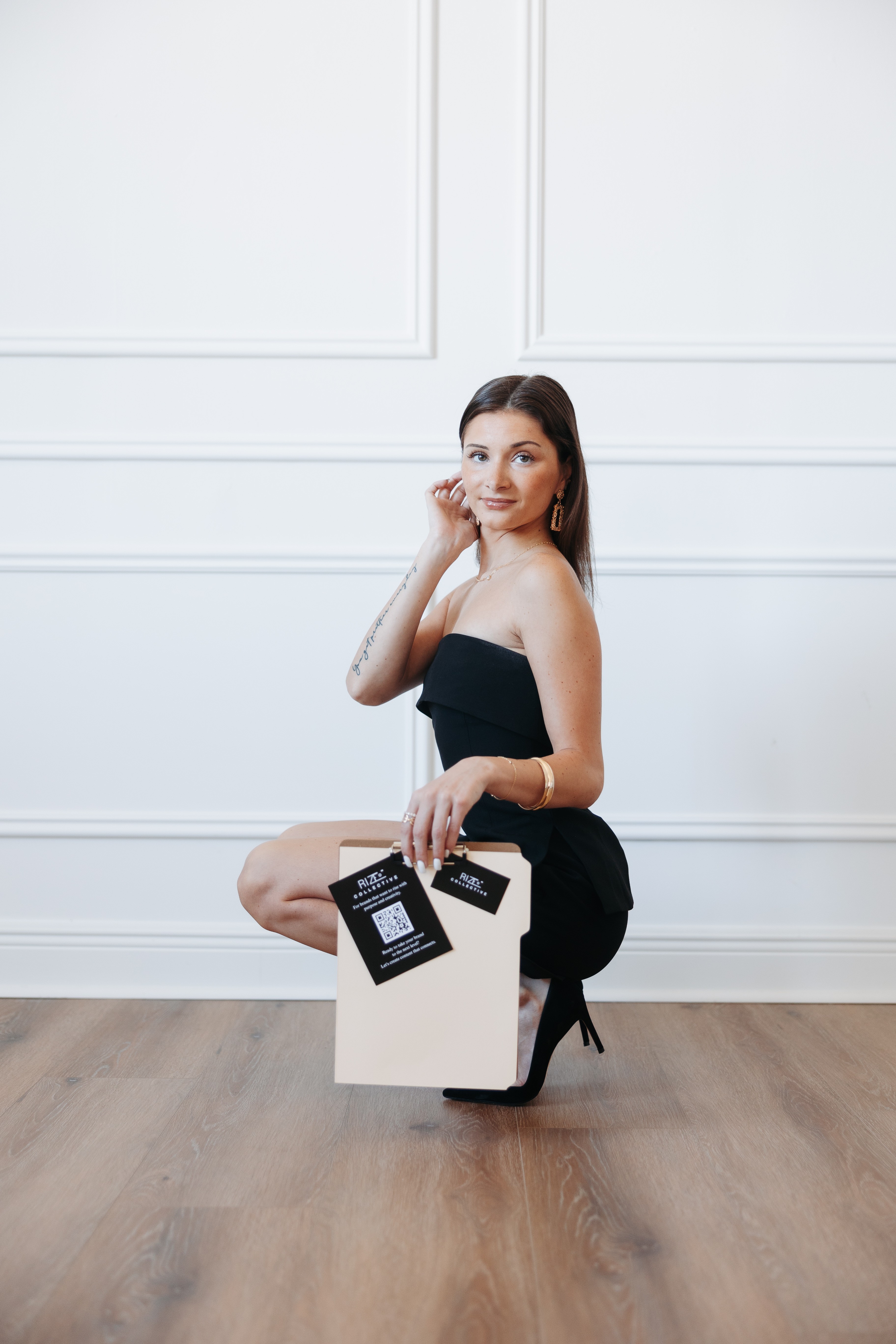 Woman in black dress sitting on a white box in a room with white walls and wooden floor, holding a card.
