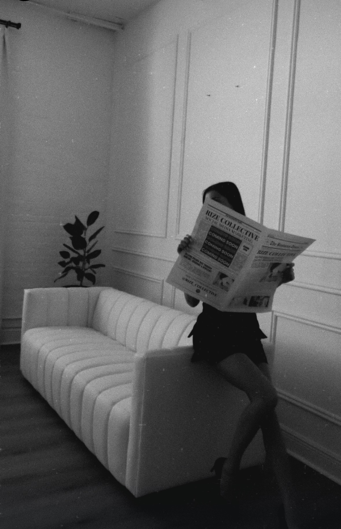 Person sitting on a sofa reading a newspaper in a room with decorative wall panels and a potted plant.