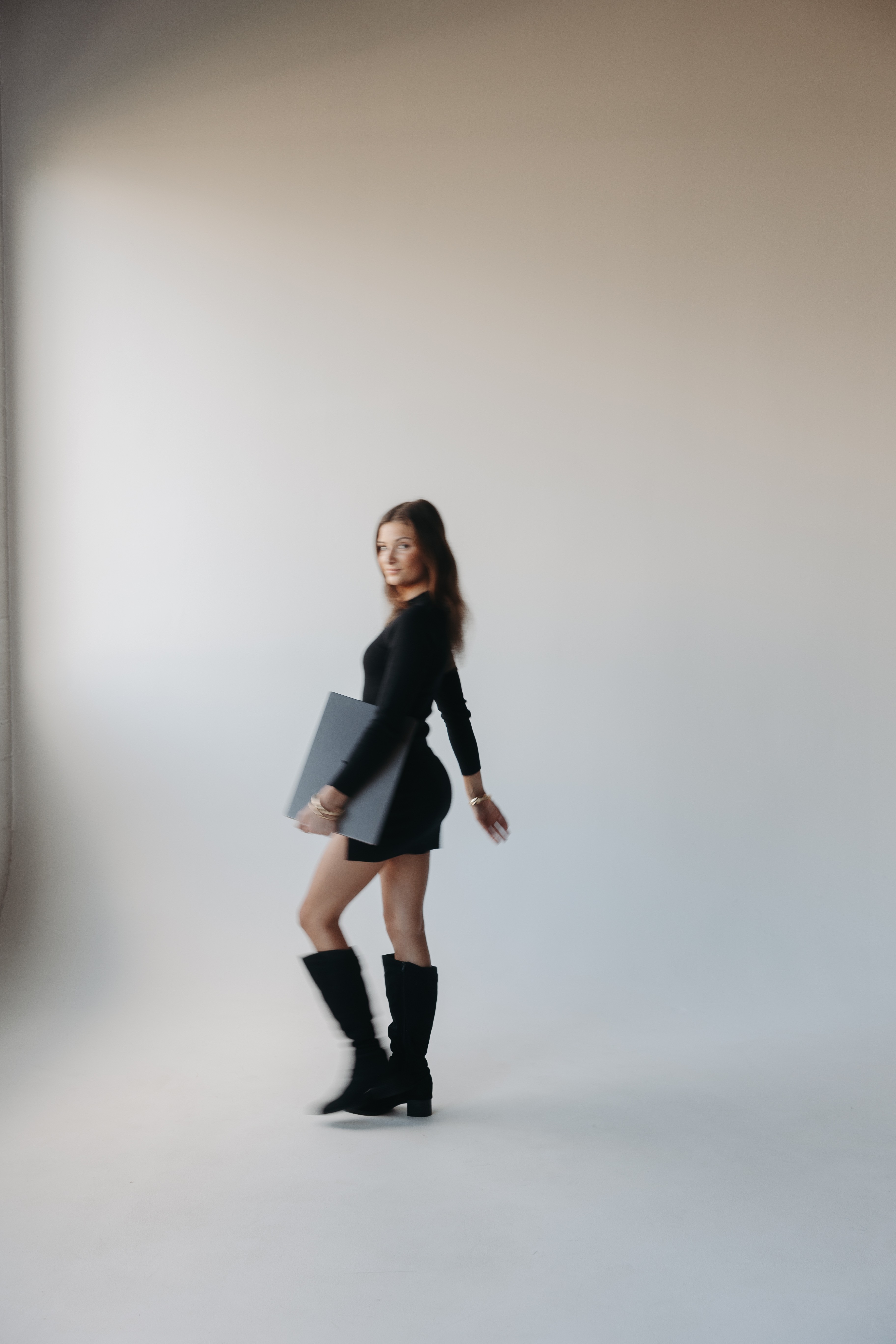 Woman standing in a studio, holding a laptop, smiling, wearing a black dress and knee-high boots.