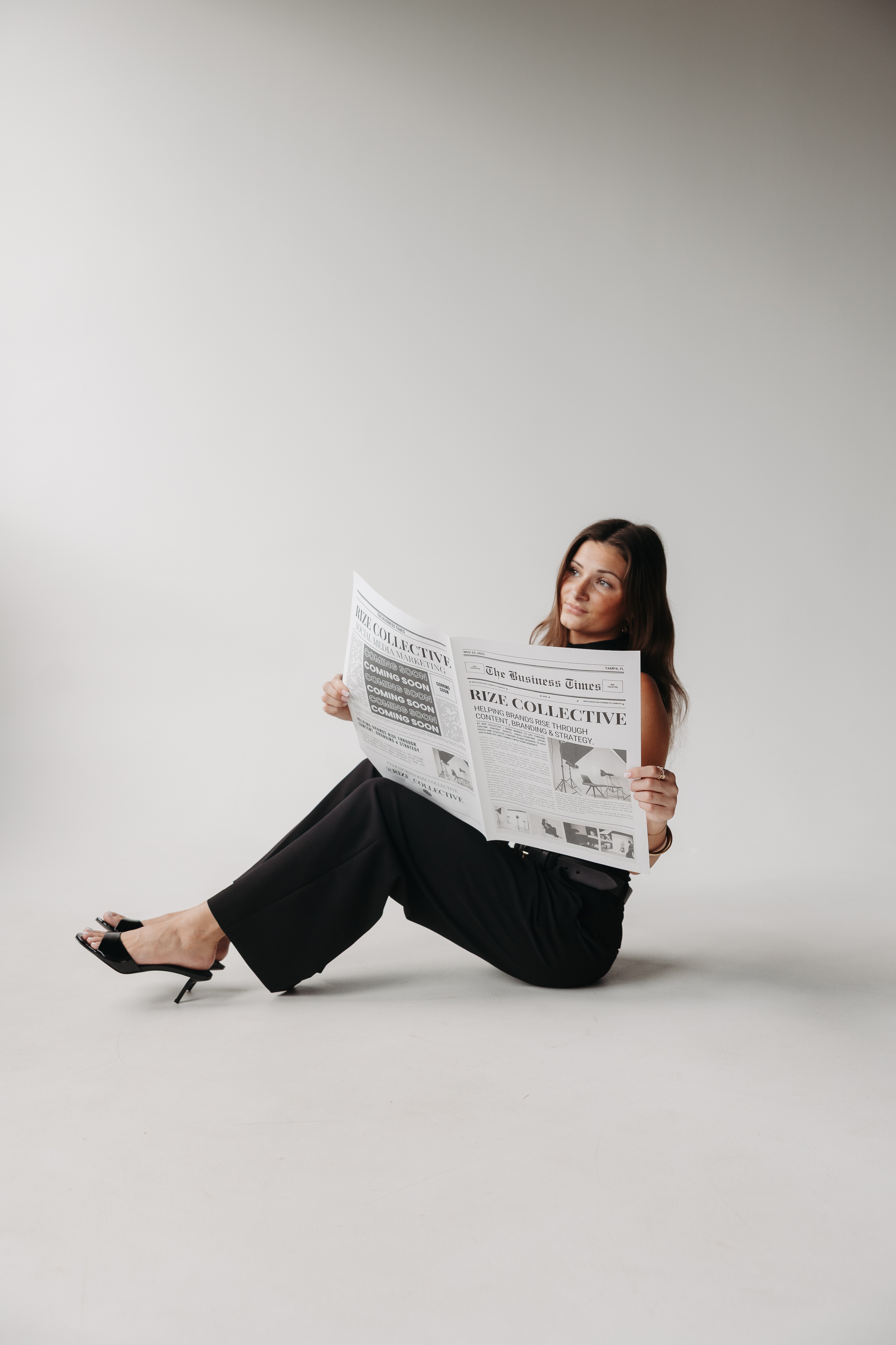 Woman sitting on the floor reading a newspaper against a plain background.