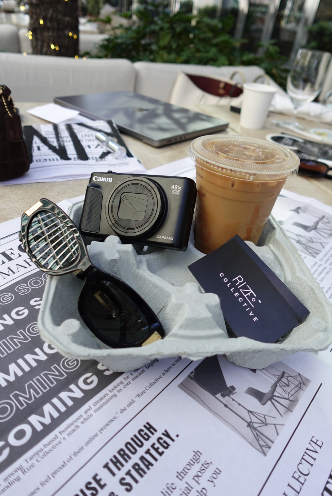 Coffee drink, camera, sunglasses, and papers on a table, with a RIZZI gift card. Items are scattered and overlapping.