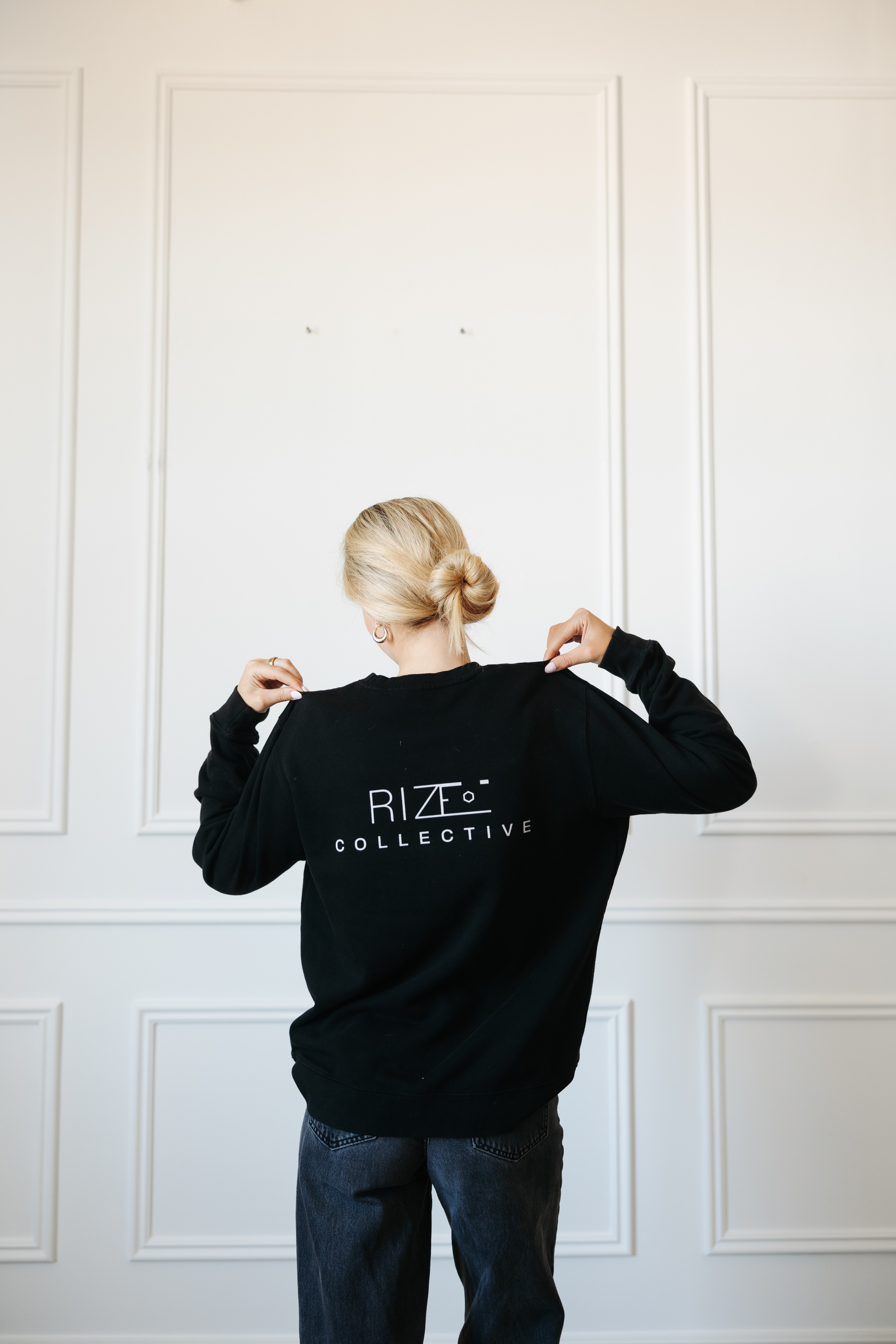 Person with blonde hair in a bun wearing a black sweatshirt with white text, facing a white wall, showing back.