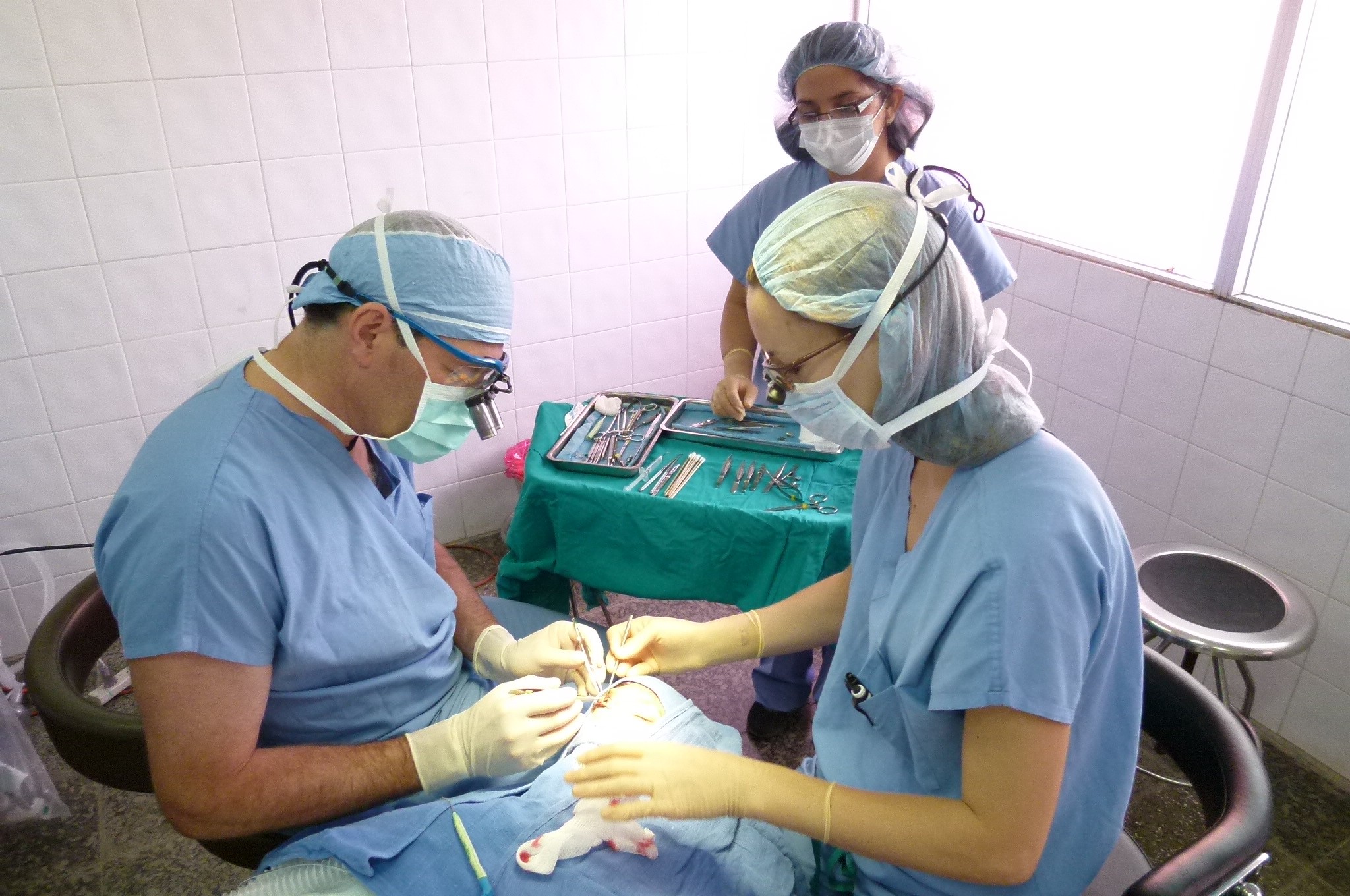 Three medical professionals in scrubs and masks perform a procedure in a clinical setting with a patient. One stands, two sit.