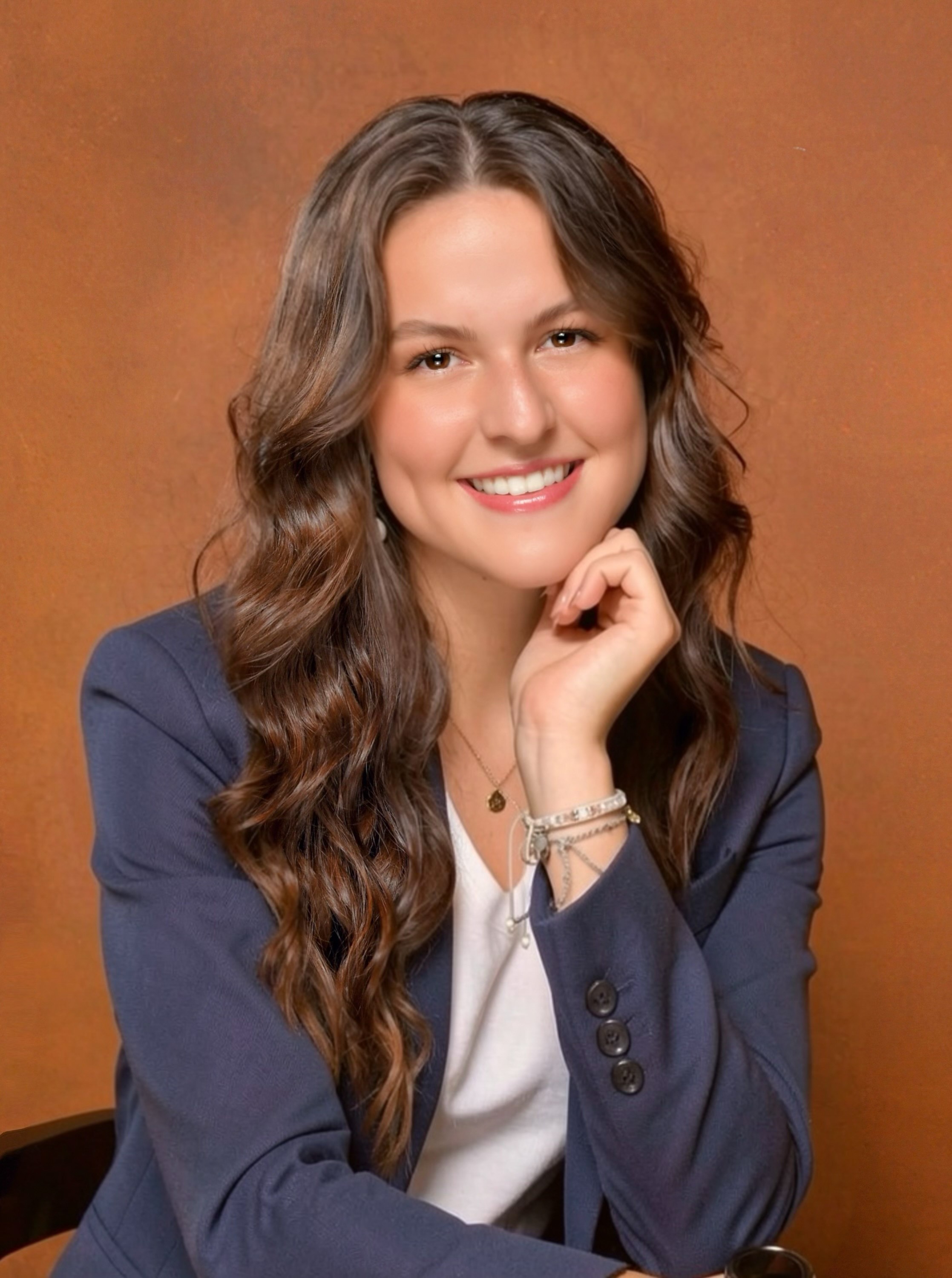 Young woman with long wavy hair smiling, wearing a navy blazer and white top, resting her chin on her hand.