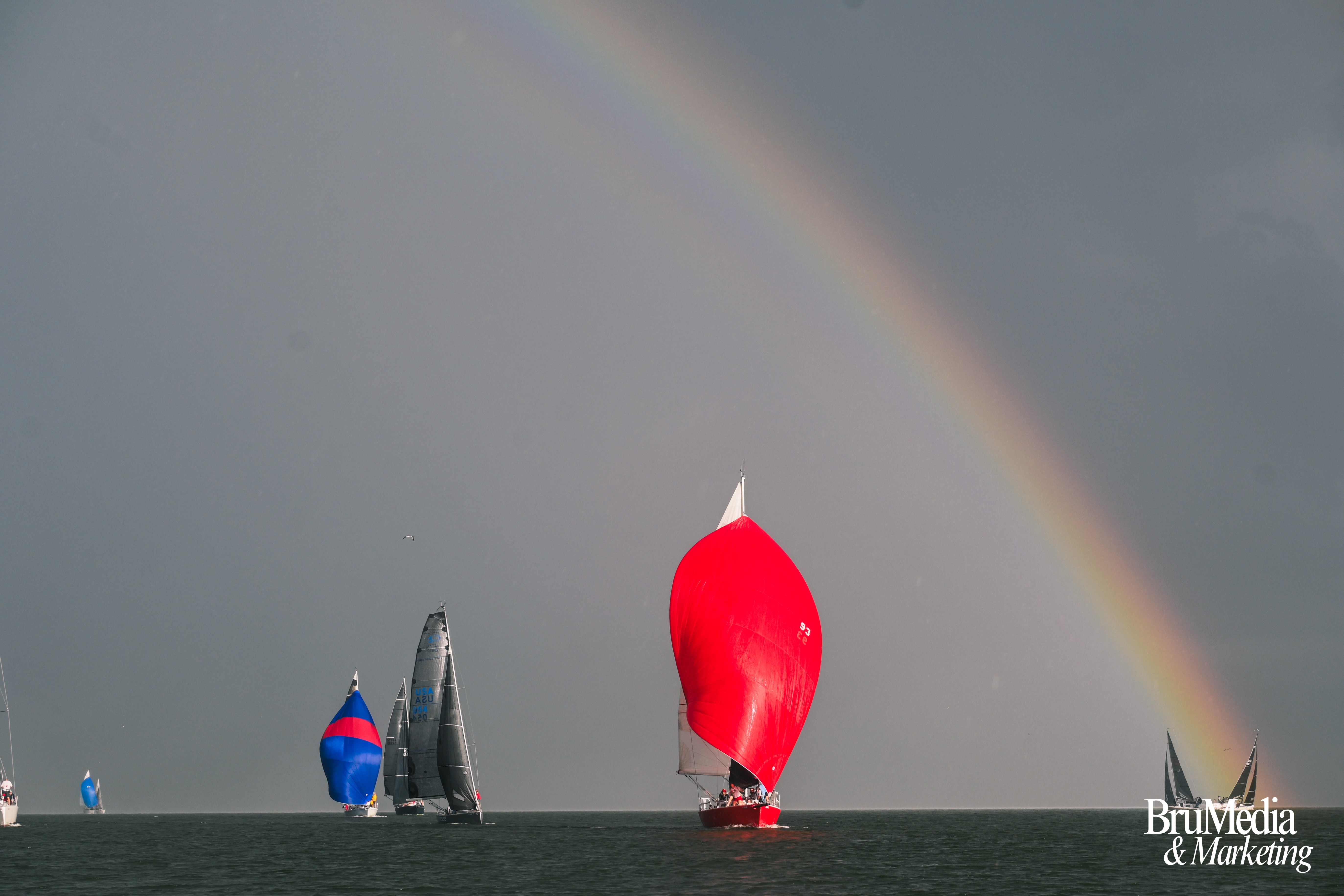 Two sailboats with colorful spinnakers on the water under a rainbow in a cloudy sky.