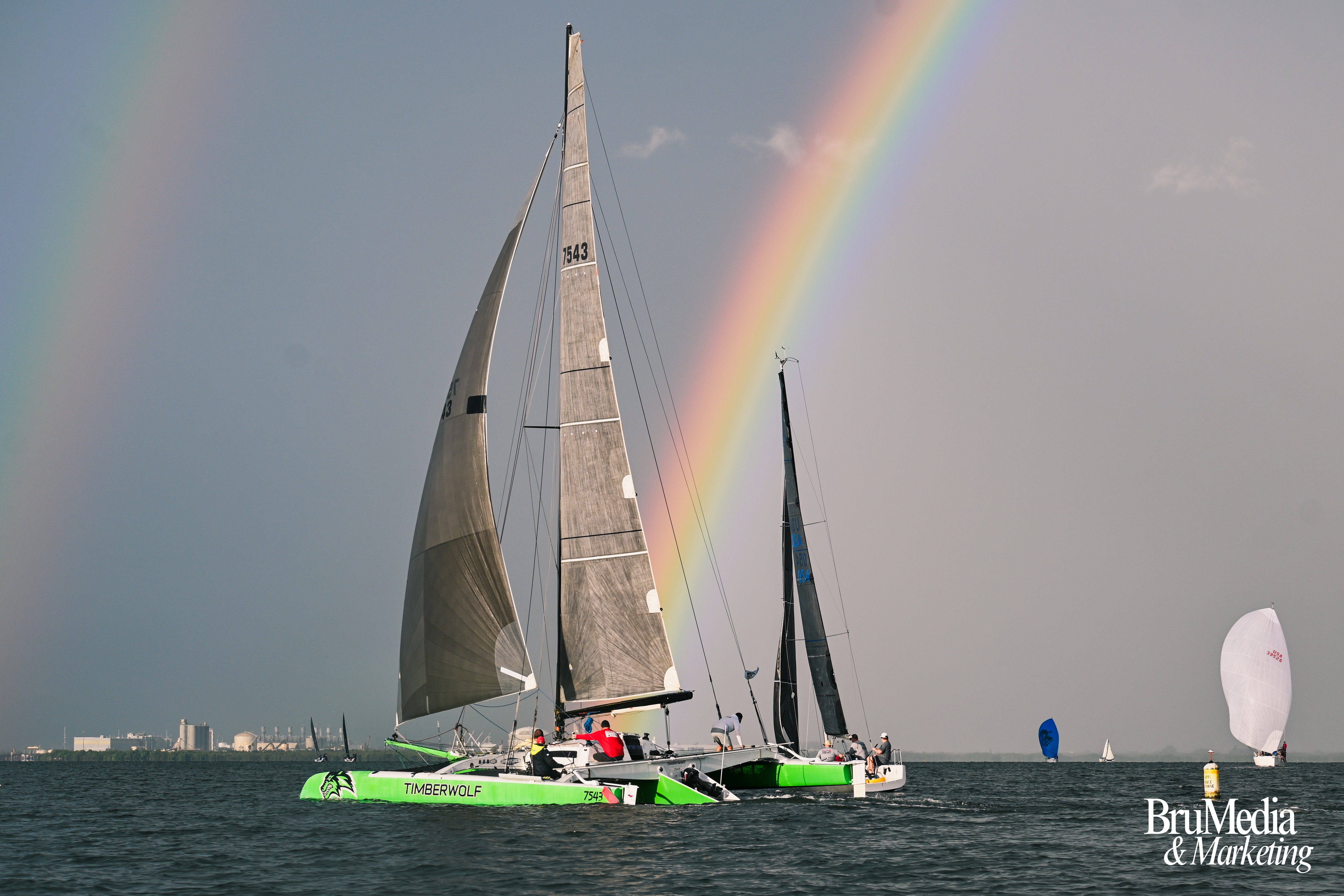 Sailboats on water with rainbow in sky, city skyline in background, cloudy weather, and a small white sailboat in distance.