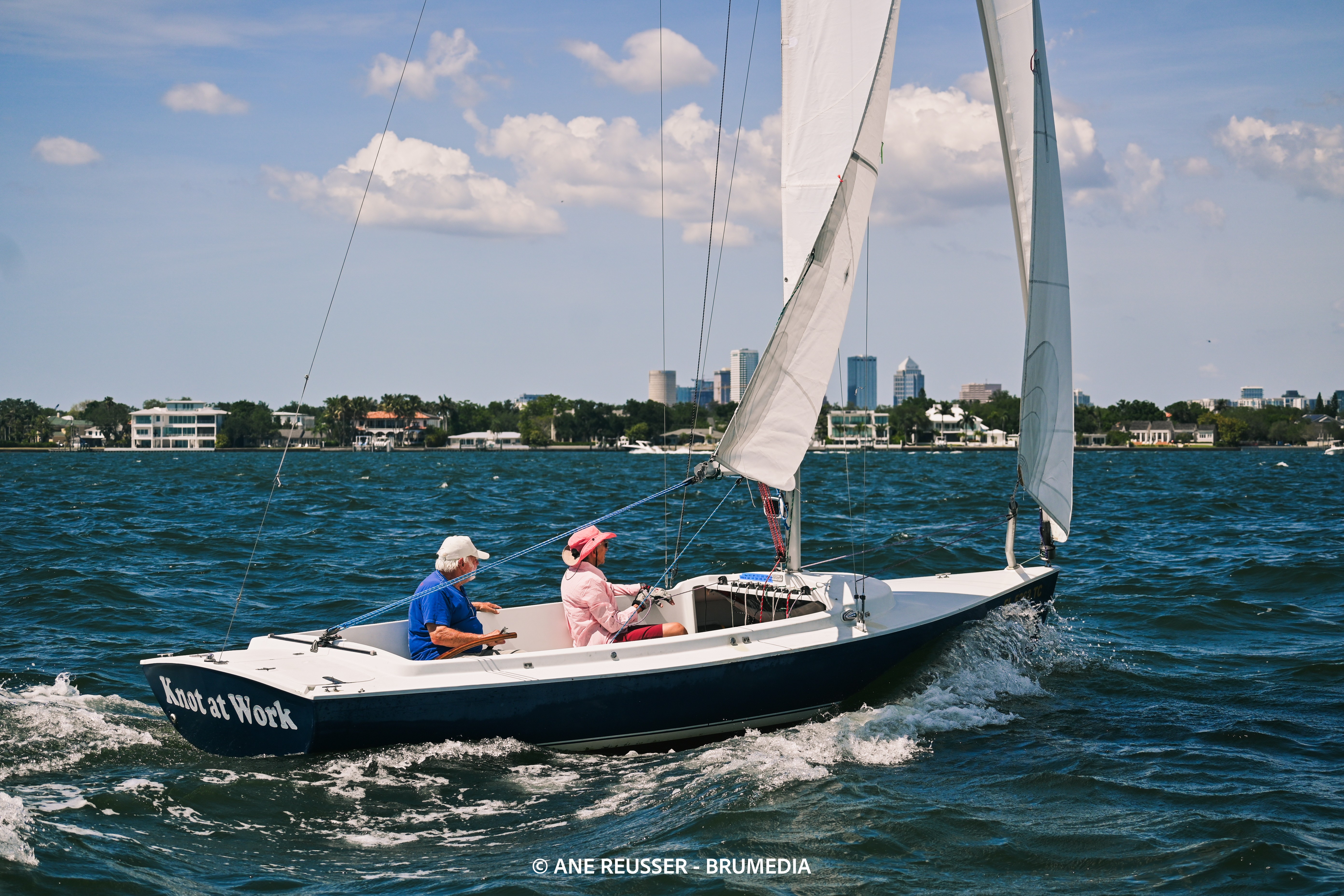 Two people sailing a boat on water with a cityscape in the background, blue sky with clouds, and the boat's sail up.