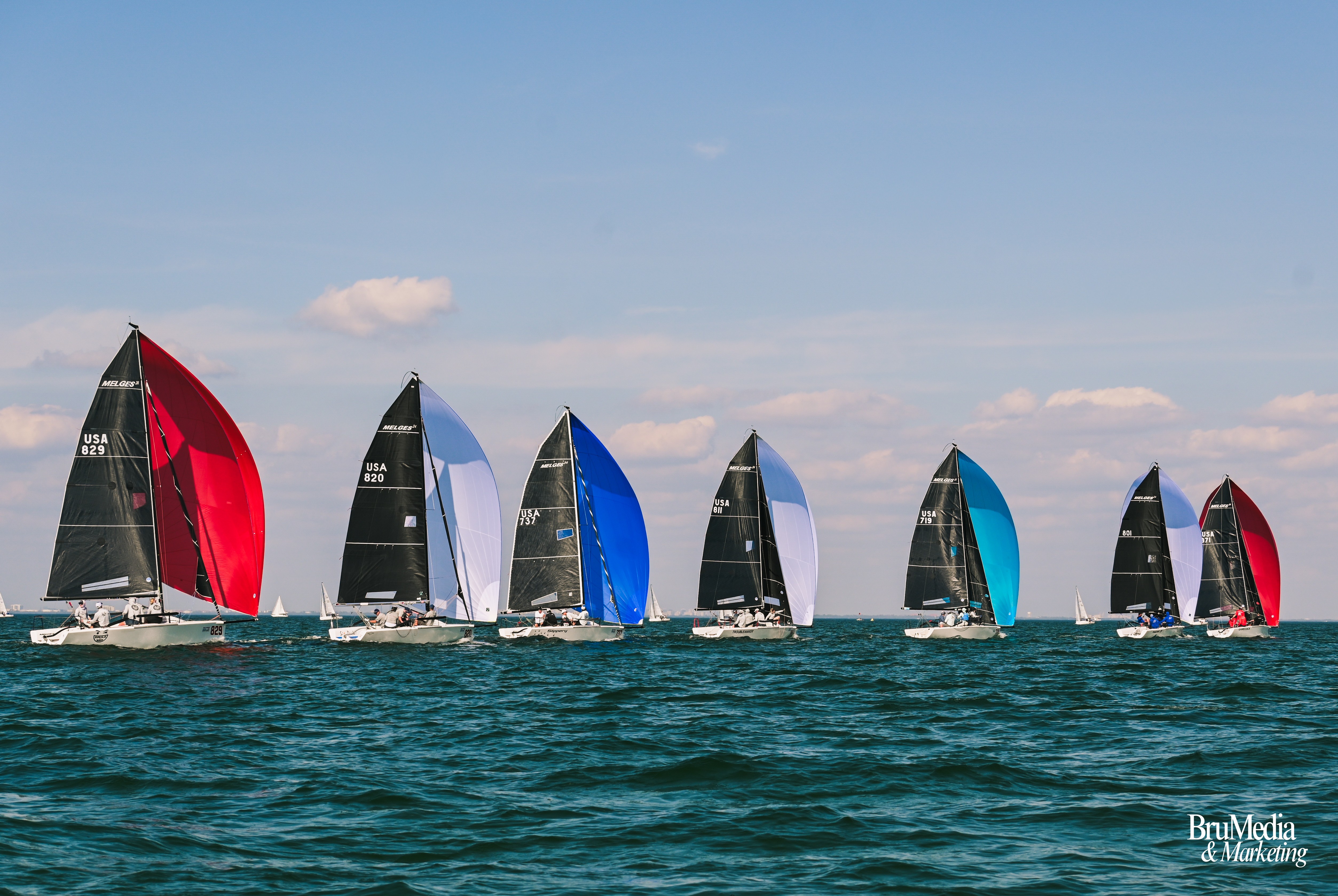 Multiple sailboats with colorful sails sailing on the water under a blue sky with clouds.