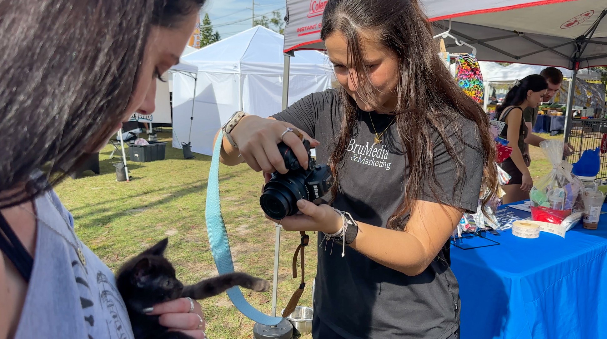 Young woman holding a camera and taking a photo of a person outdoors at an event with tents and tables.