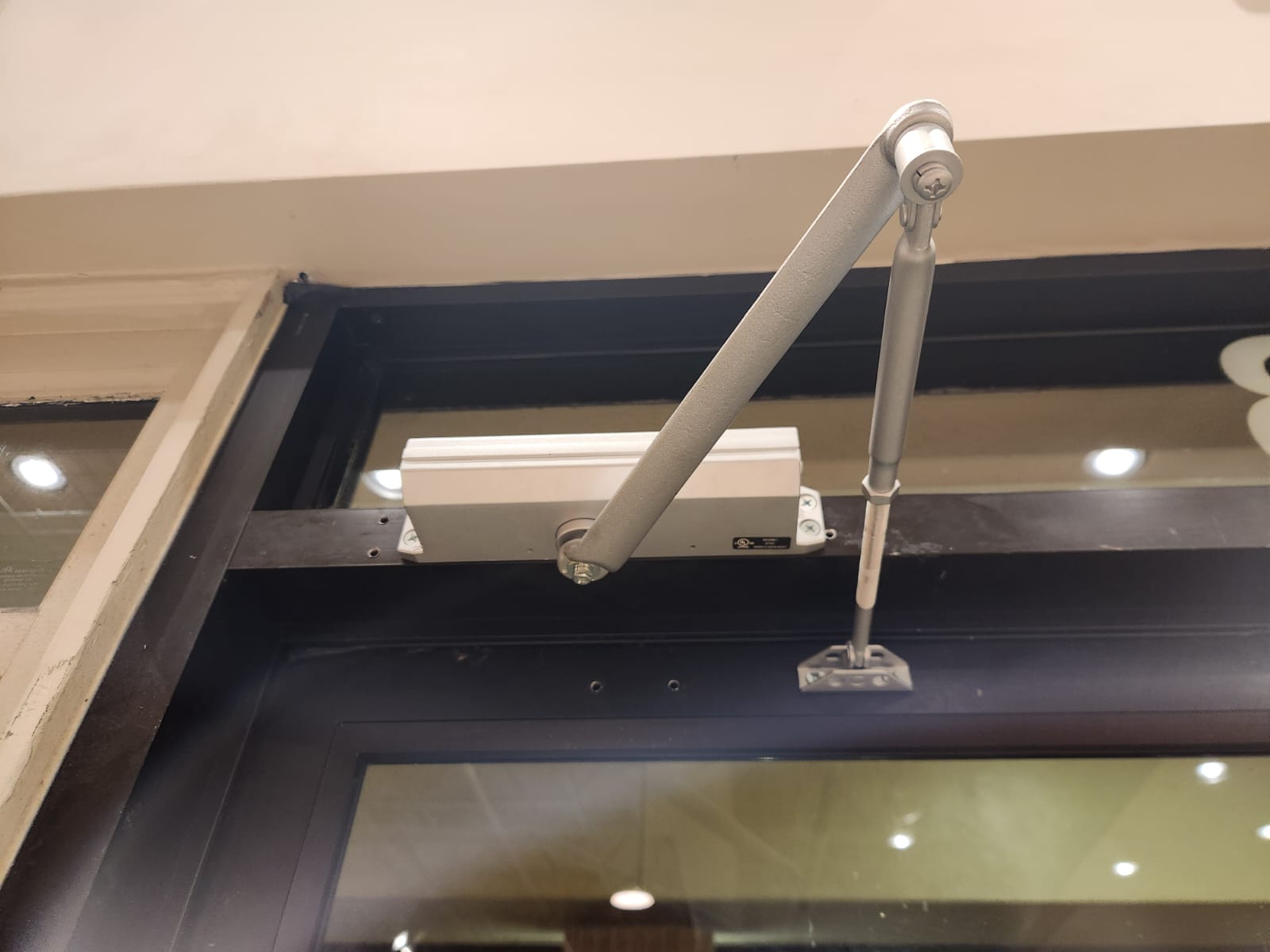 Close-up of a door frame with a metal arm mechanism attached, part of a door closer system, with a black frame and white wall.