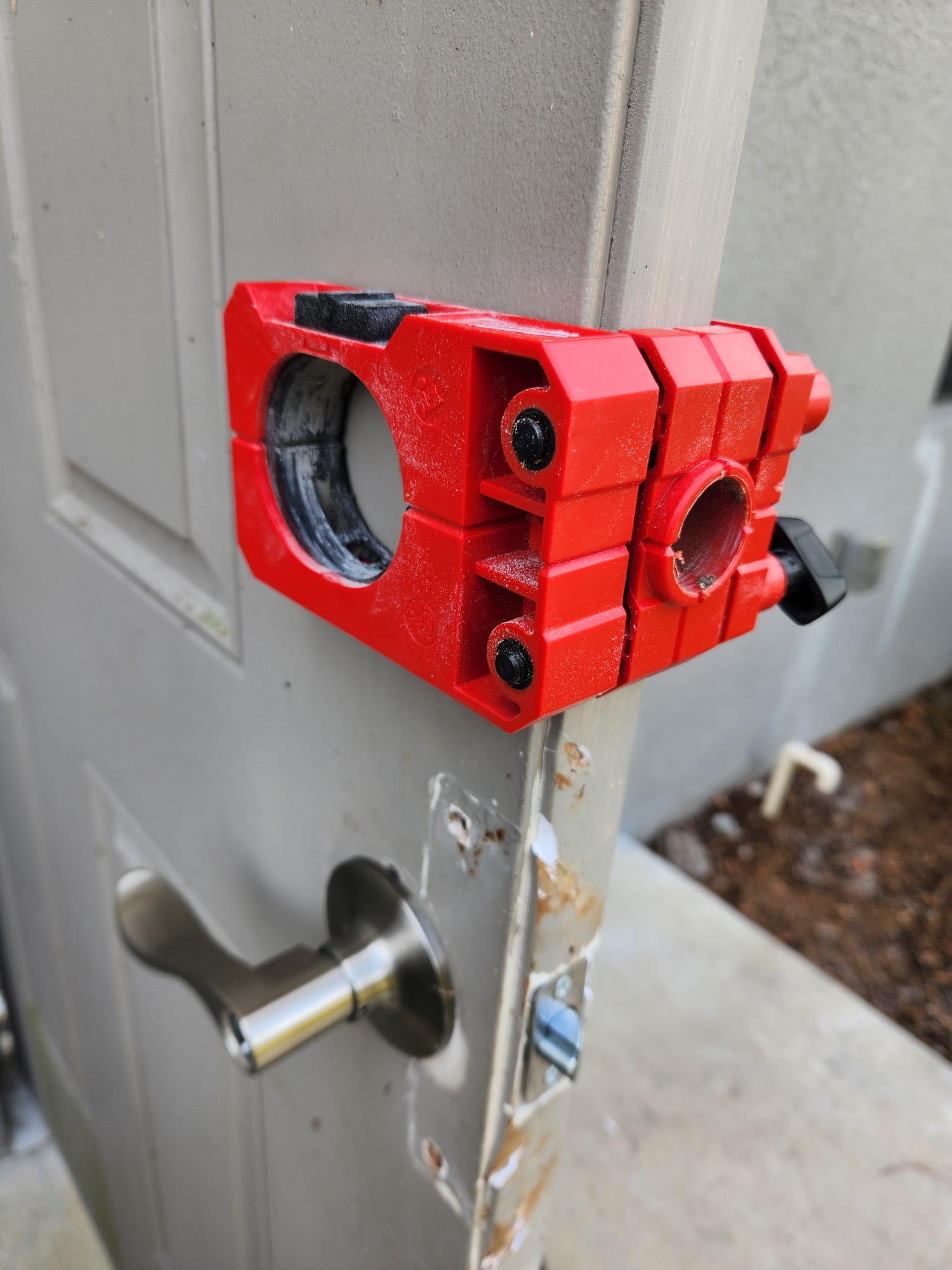 Red device attached to a gray door, with a circular opening and a black knob, outdoors.
