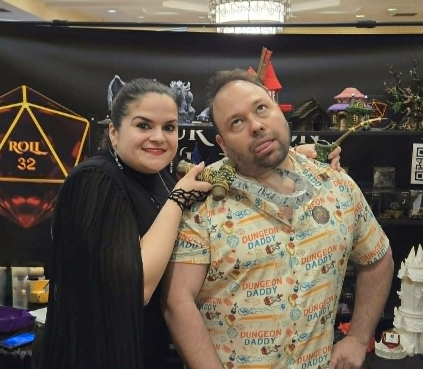 A woman with dark hair and a man with short hair pose together indoors, with a black backdrop and decorations behind them.