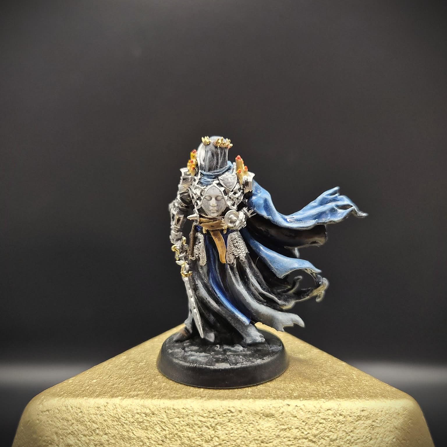 Miniature figure of a person in elaborate clothing with flowing fabric, standing on a round base, on a textured surface.