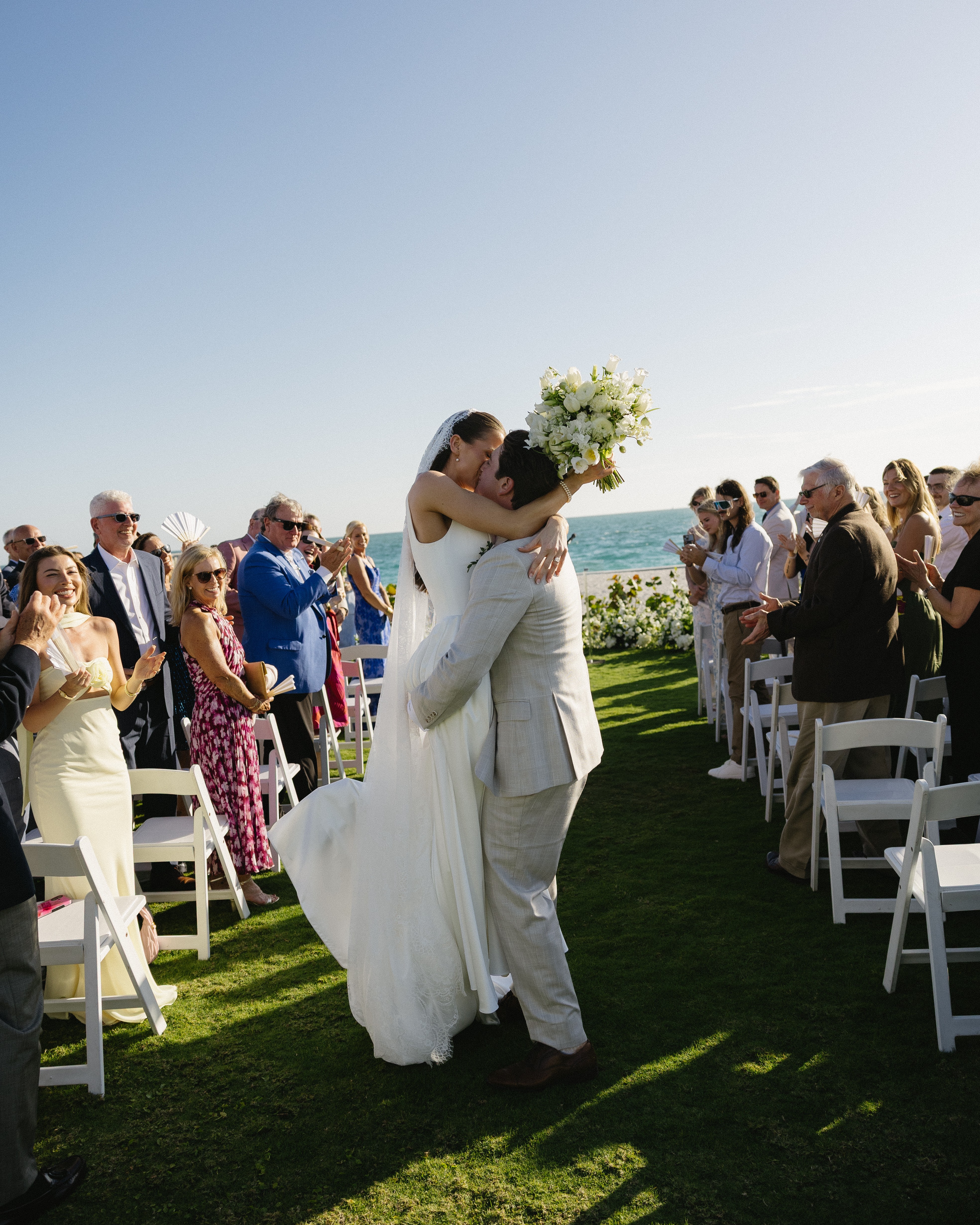 A bride and groom kiss outdoors near the ocean, surrounded by seated guests, with a bouquet of flowers, under a clear sky.