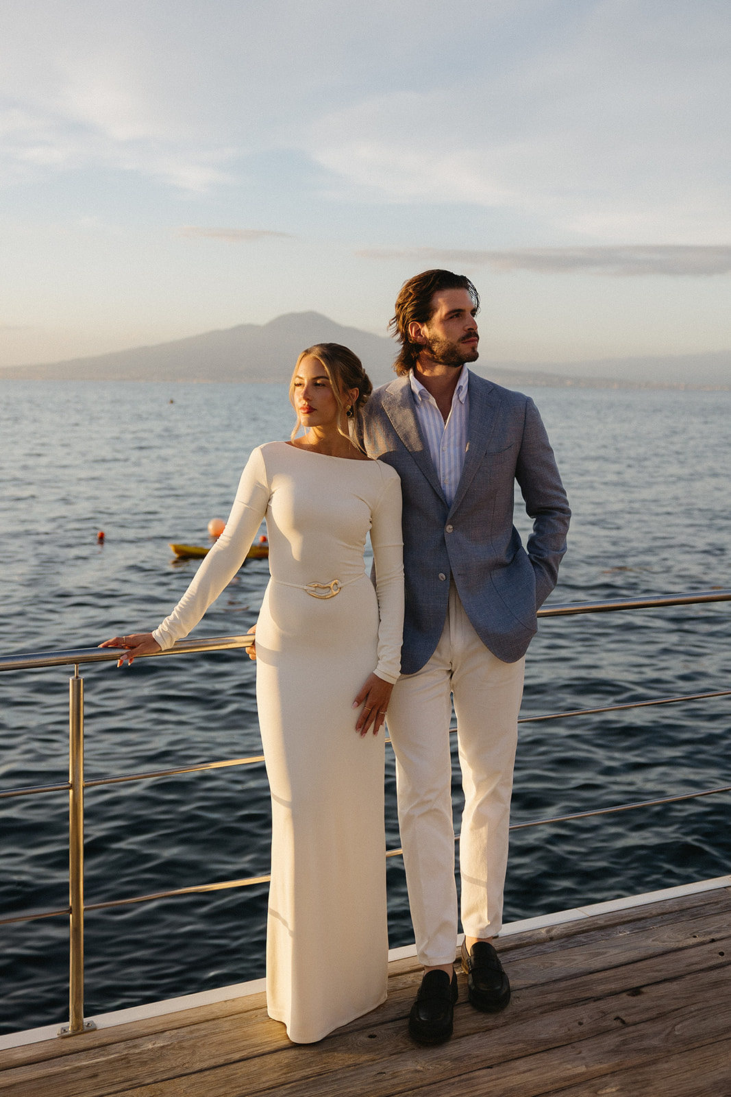 A man and woman stand on a dock by the water, with mountains and sky in the background, dressed elegantly.