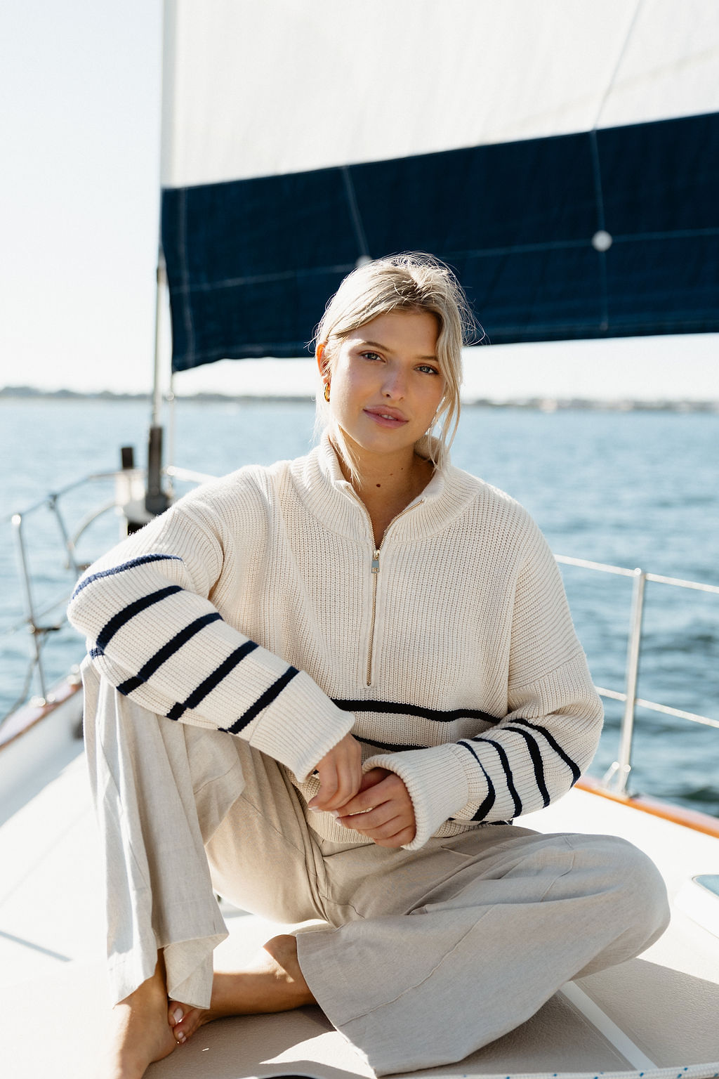 Woman sitting on a boat deck with water and sail in background, wearing a cream sweater with black stripes.