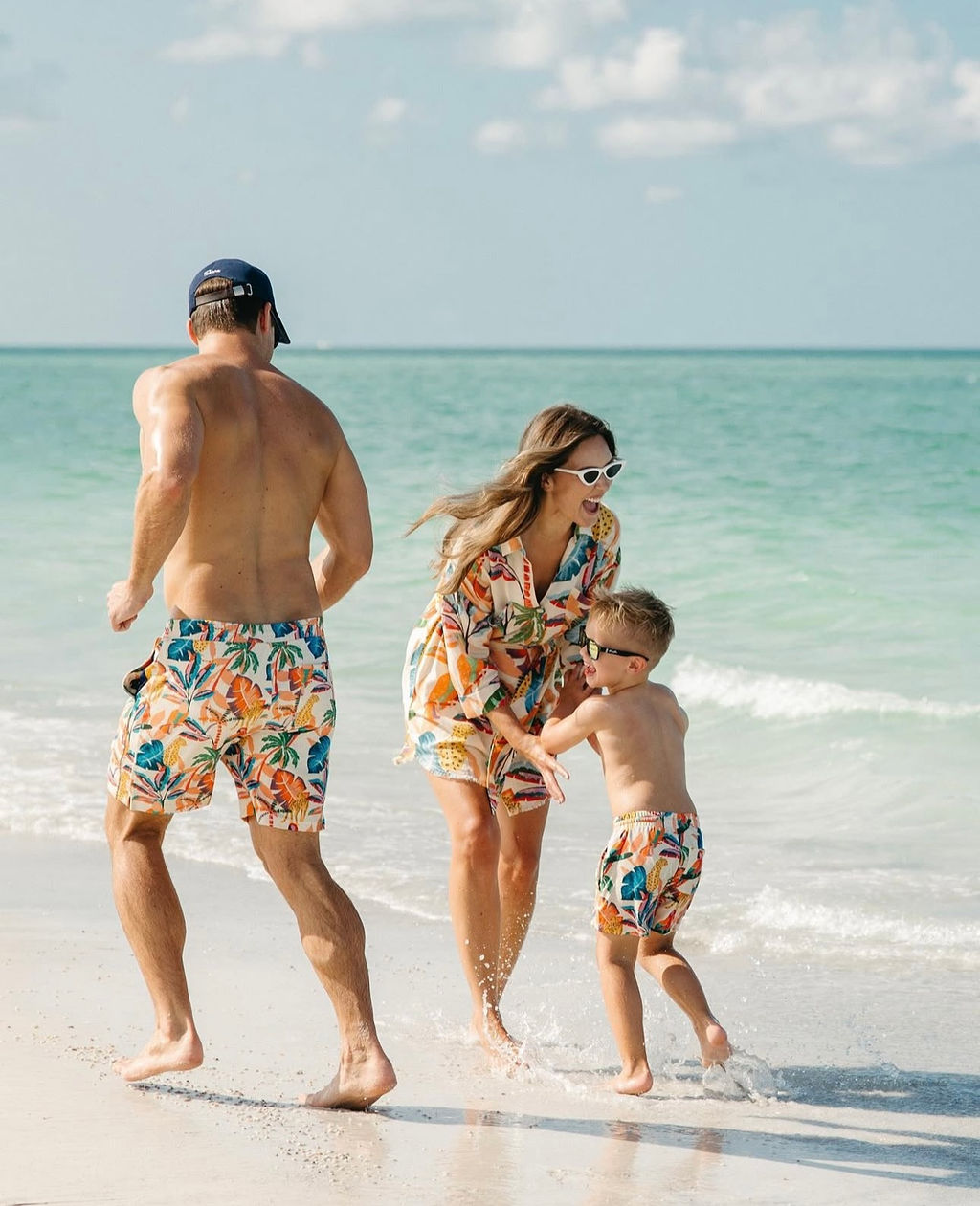 Three people, two adults and a child, on a beach near the water, wearing colorful swimwear and sunglasses, enjoying the day.