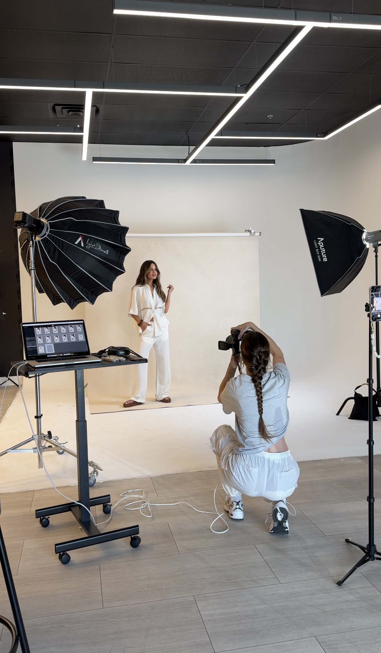 Photo shoot setup with a model in white clothing, a photographer crouching, and studio lighting equipment in a modern studio.