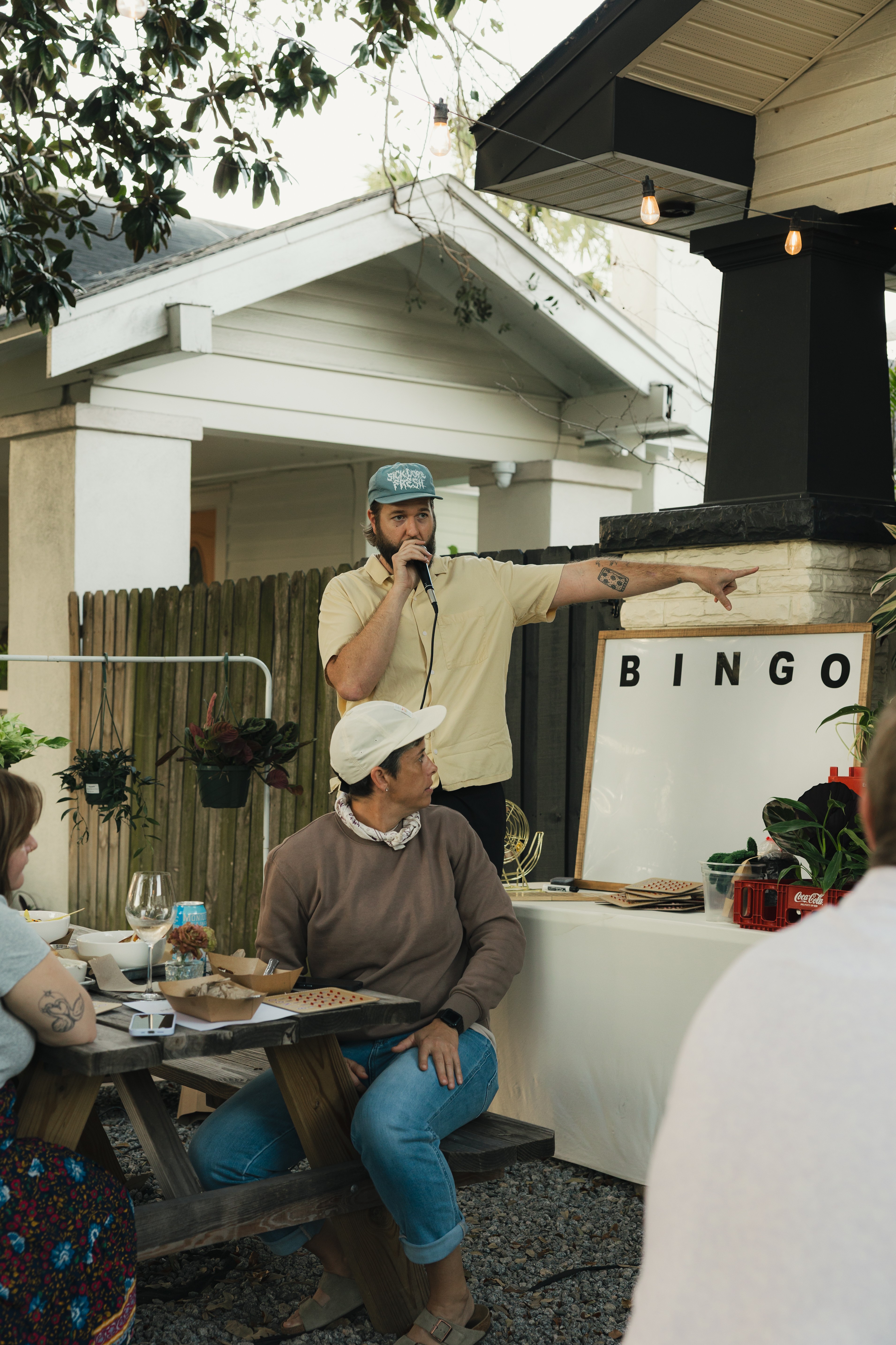 Man with a beard and cap points at a bingo game, woman in white hat sits nearby, outdoor setting with house and trees