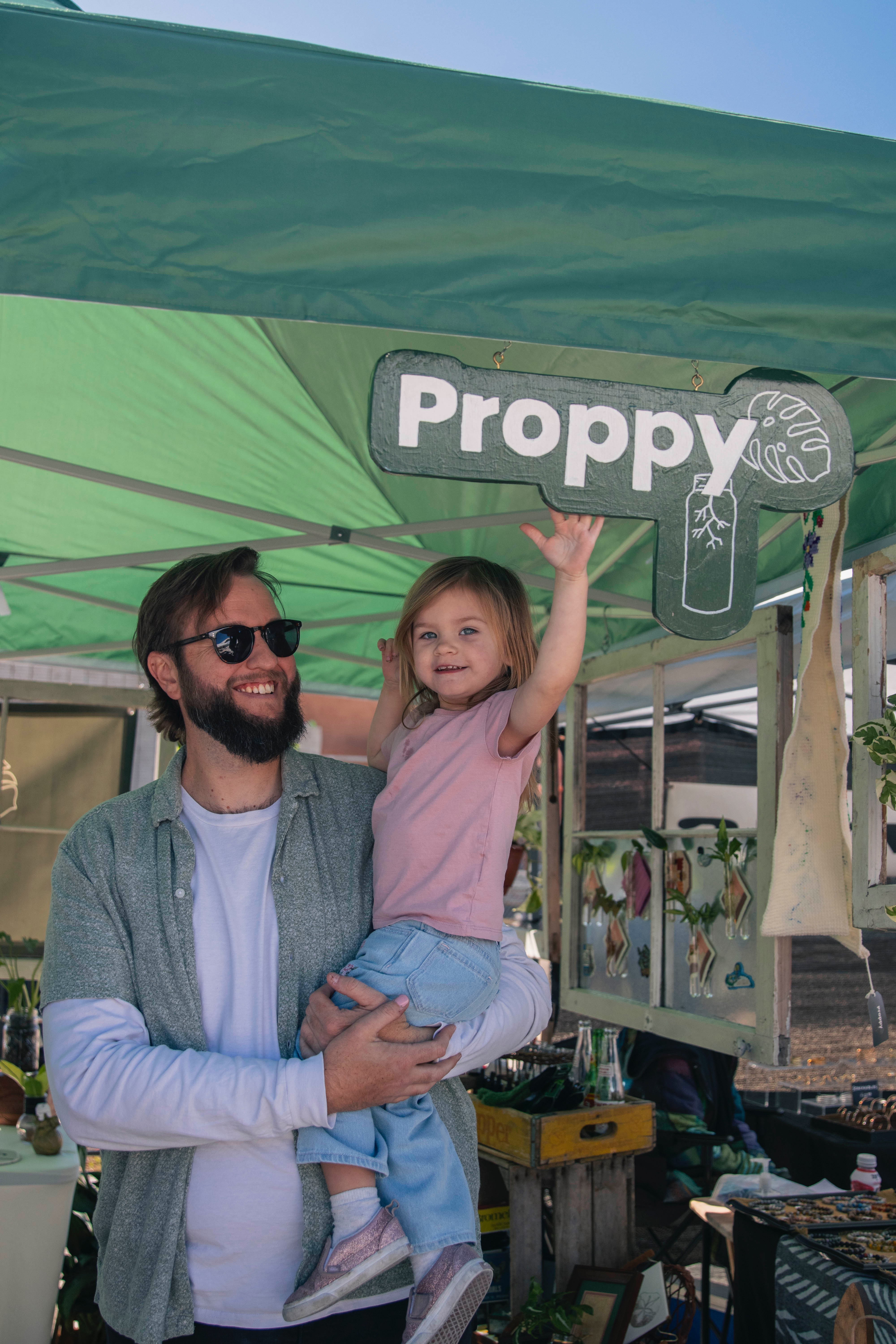Man with sunglasses holding a young girl under a green canopy at an outdoor event.