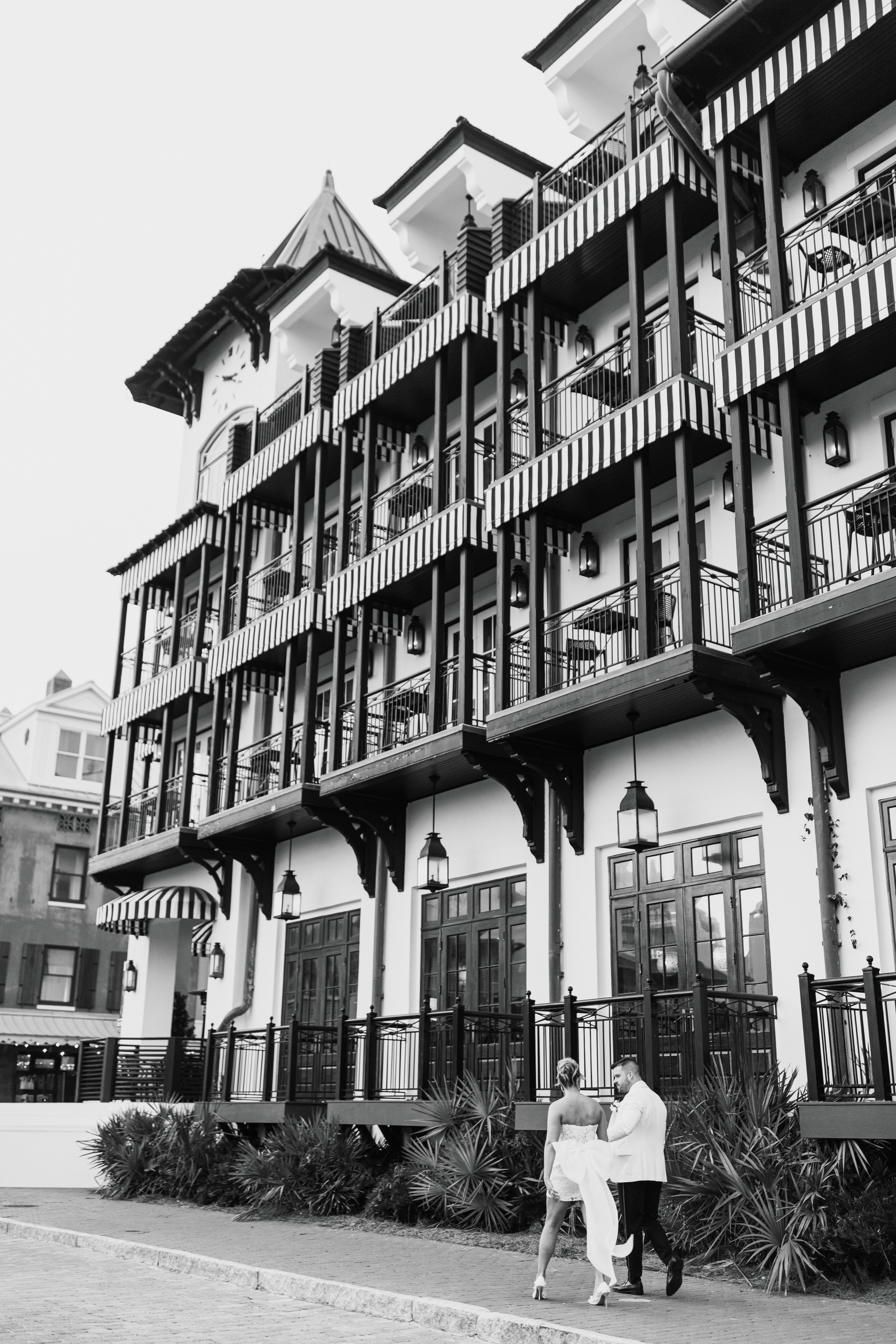 Multi-story building with balconies, outdoor seating, and two people walking in front, black and white photo.