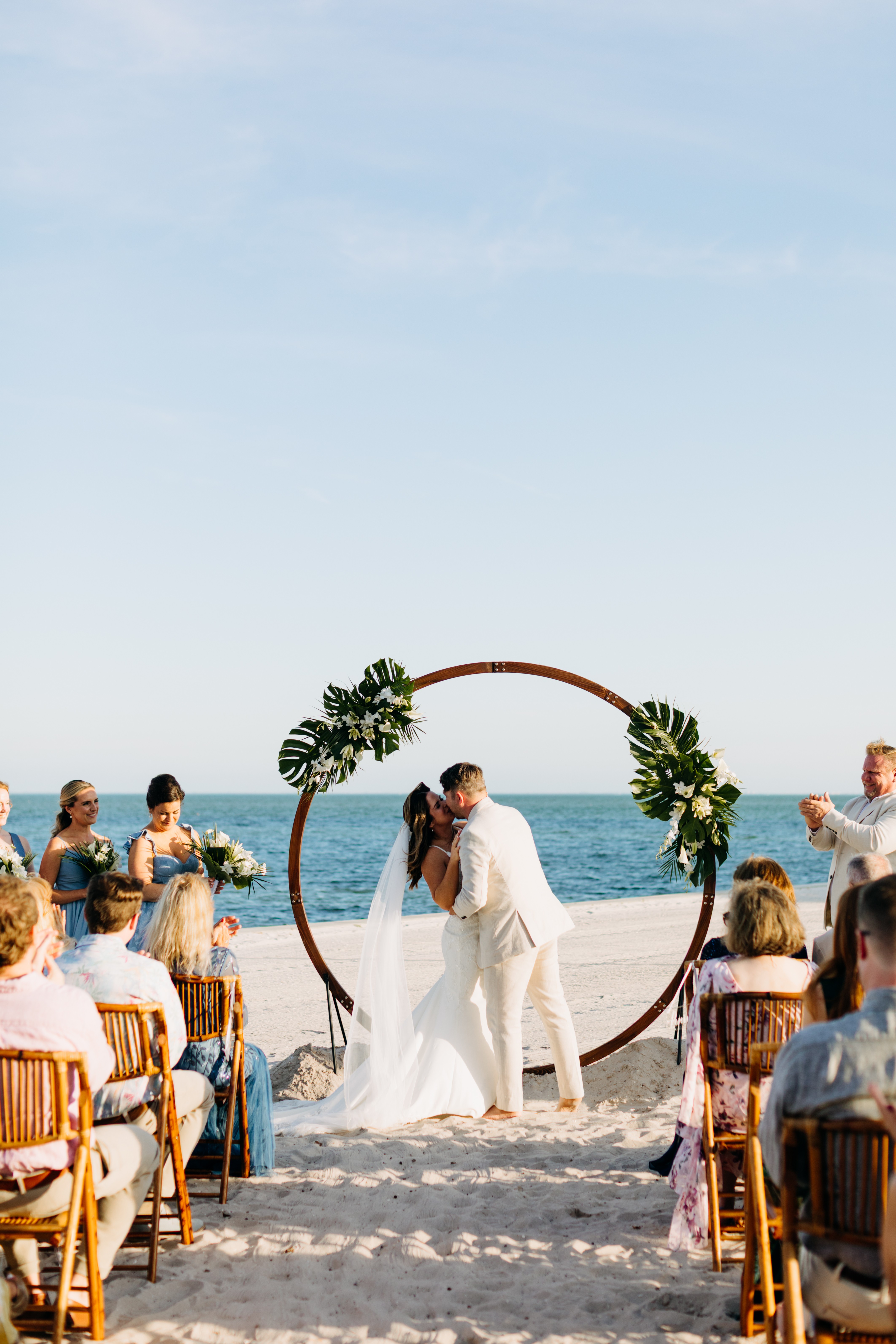 Couple kissing at beach wedding ceremony with guests seated on sand, ocean in background, decorated circular arch with greenery.