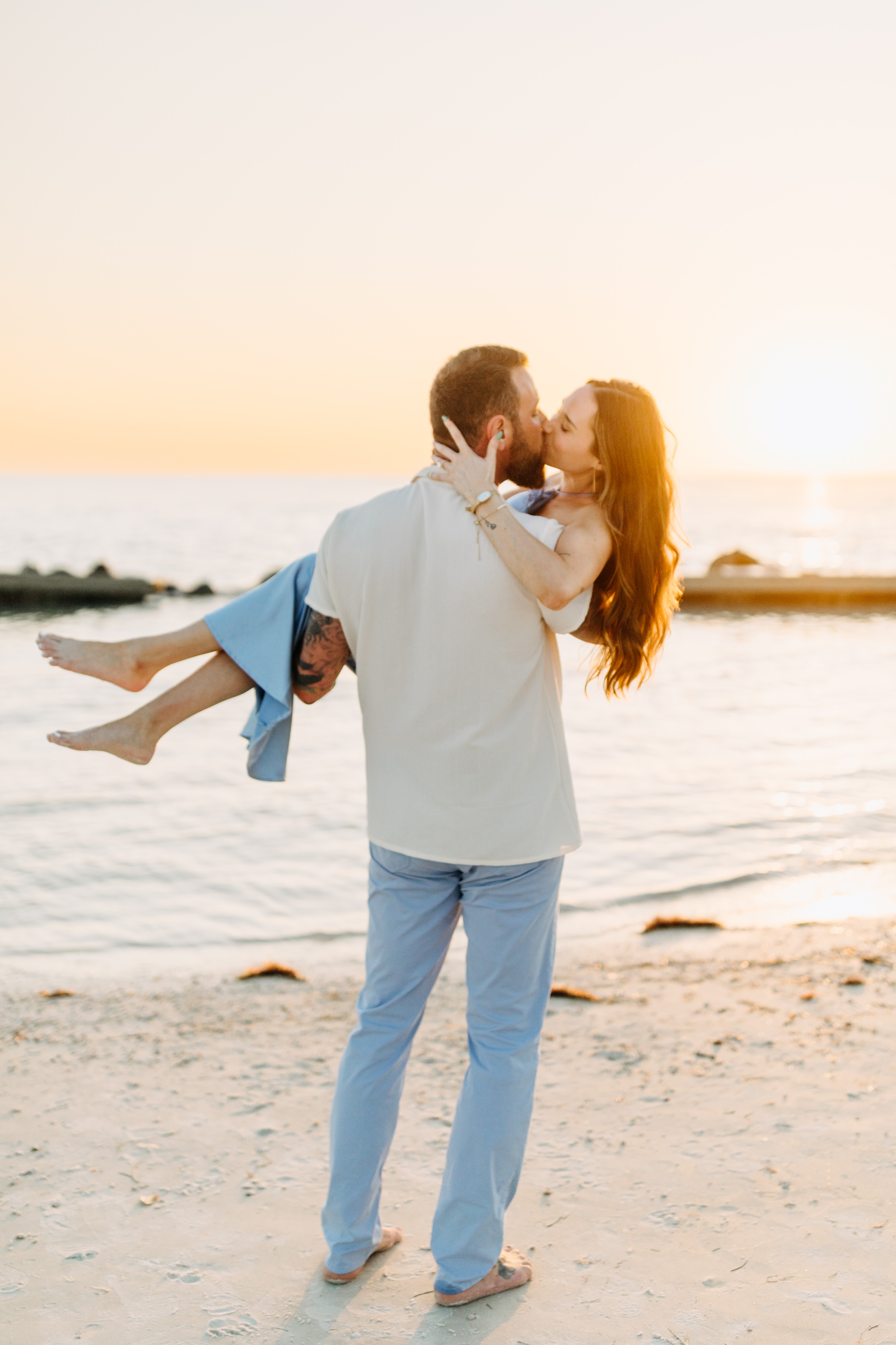 Man holding woman at the beach during sunset, both smiling, woman with long hair, man in light clothing, water in background