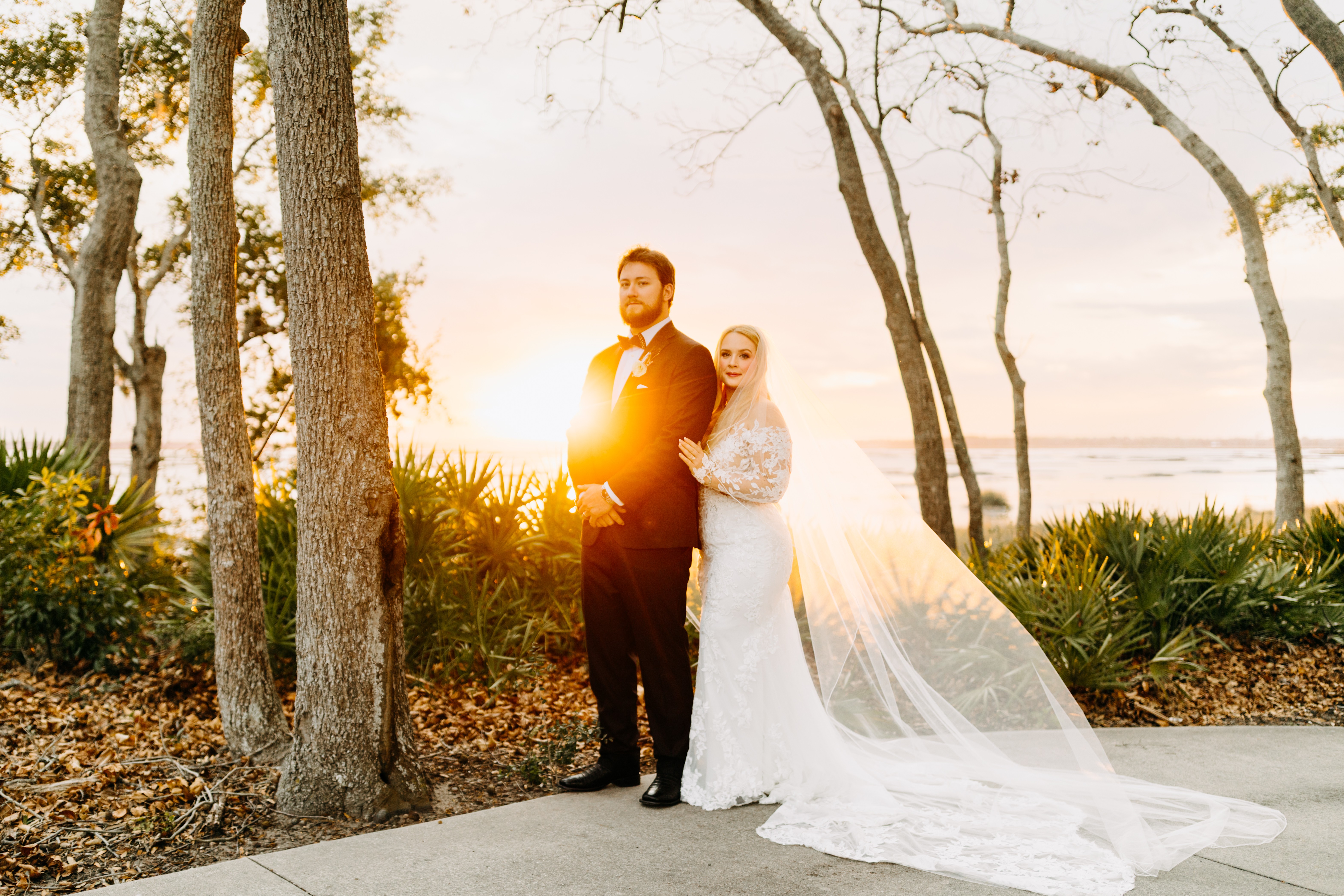 A couple in wedding attire standing outdoors near trees during sunset, with the bride's long veil flowing behind her.