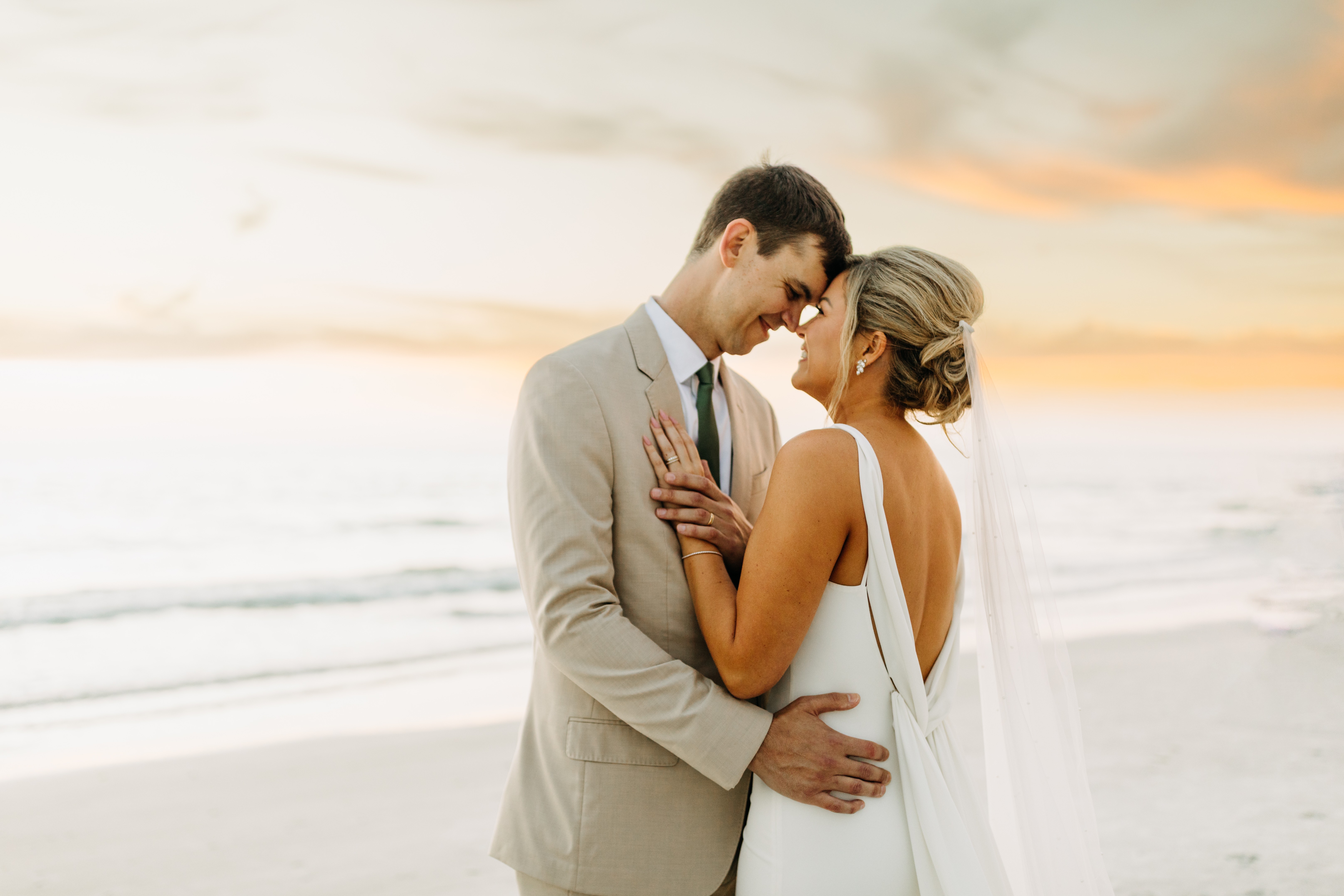 A couple in wedding attire stands close on a beach at sunset, touching foreheads and holding each other.