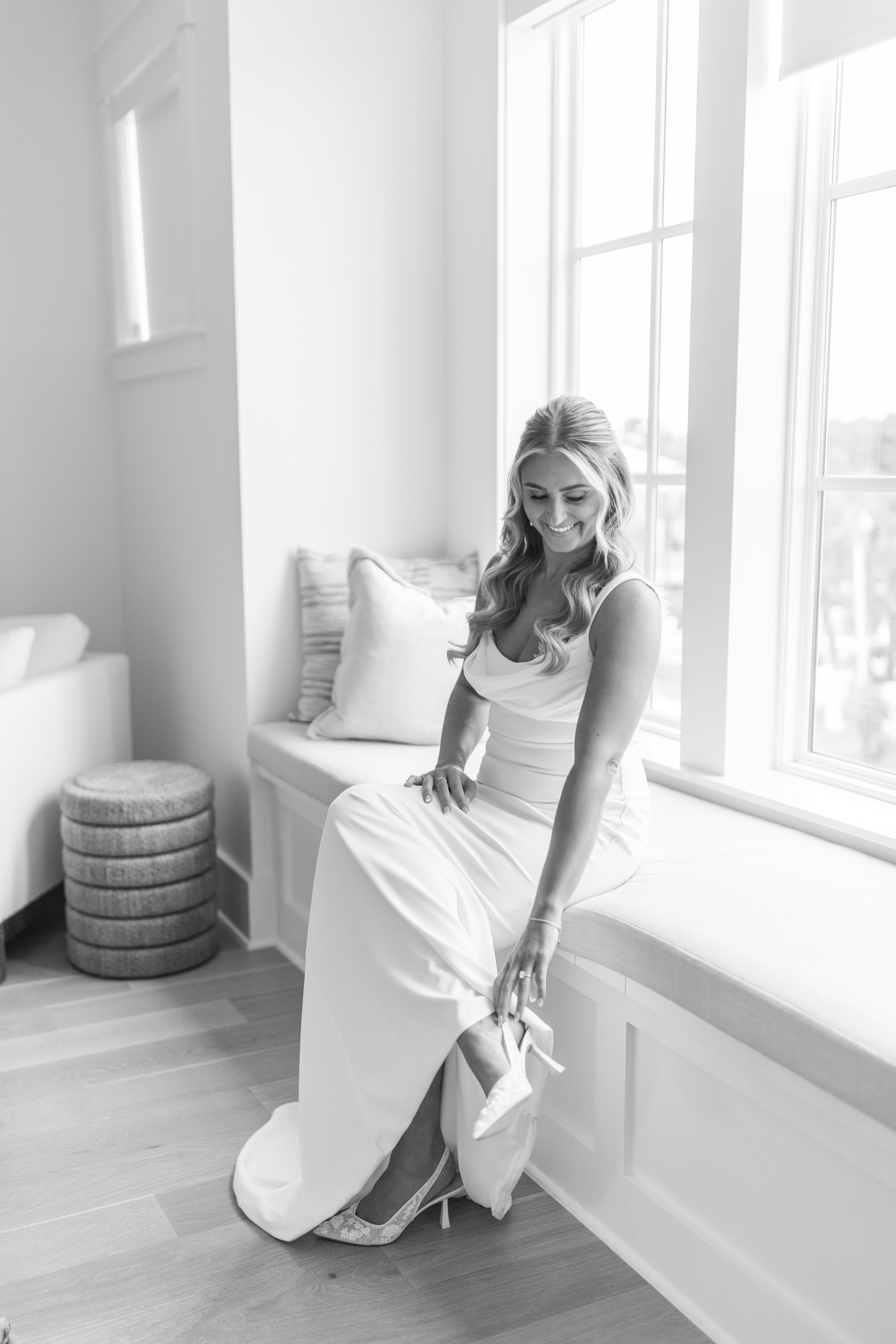 Woman in a white dress sitting by a large window, holding her shoe, with sunlight streaming in, in a bright room.
