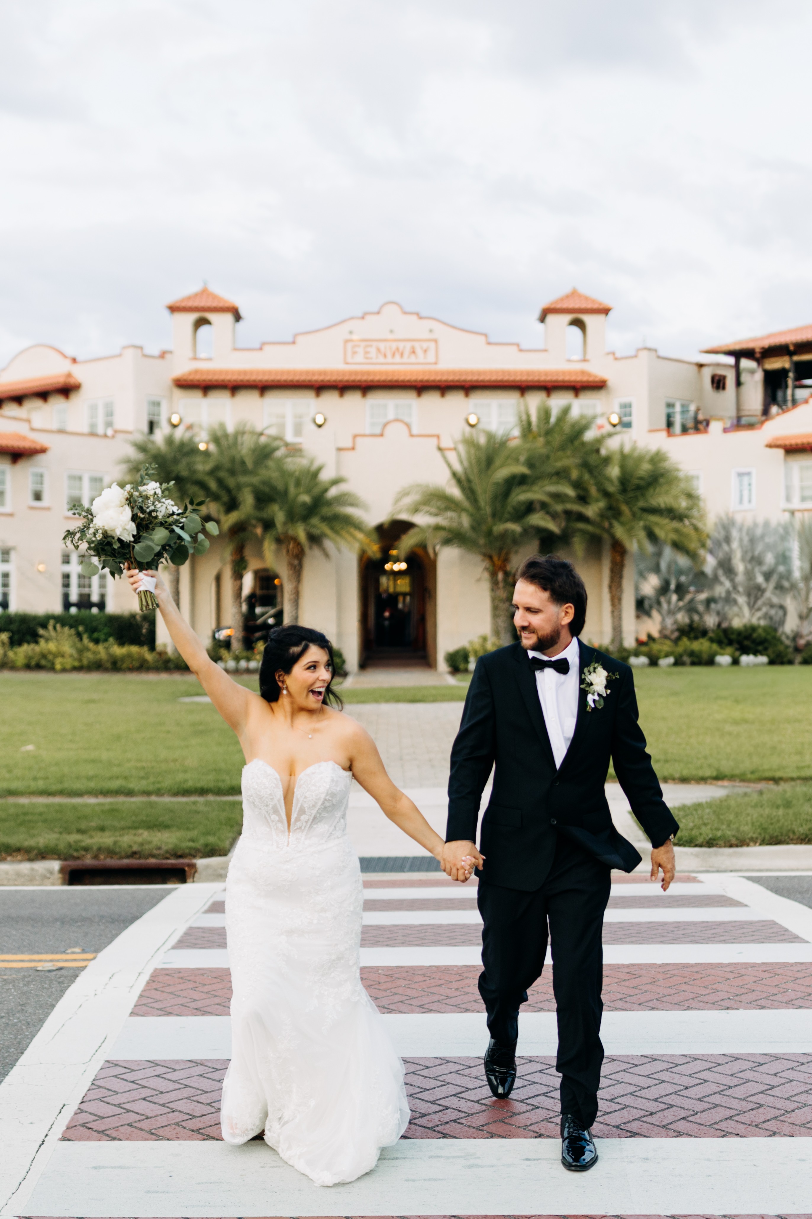 Bride and groom holding hands and walking on a crosswalk in front of a large building with palm trees.
