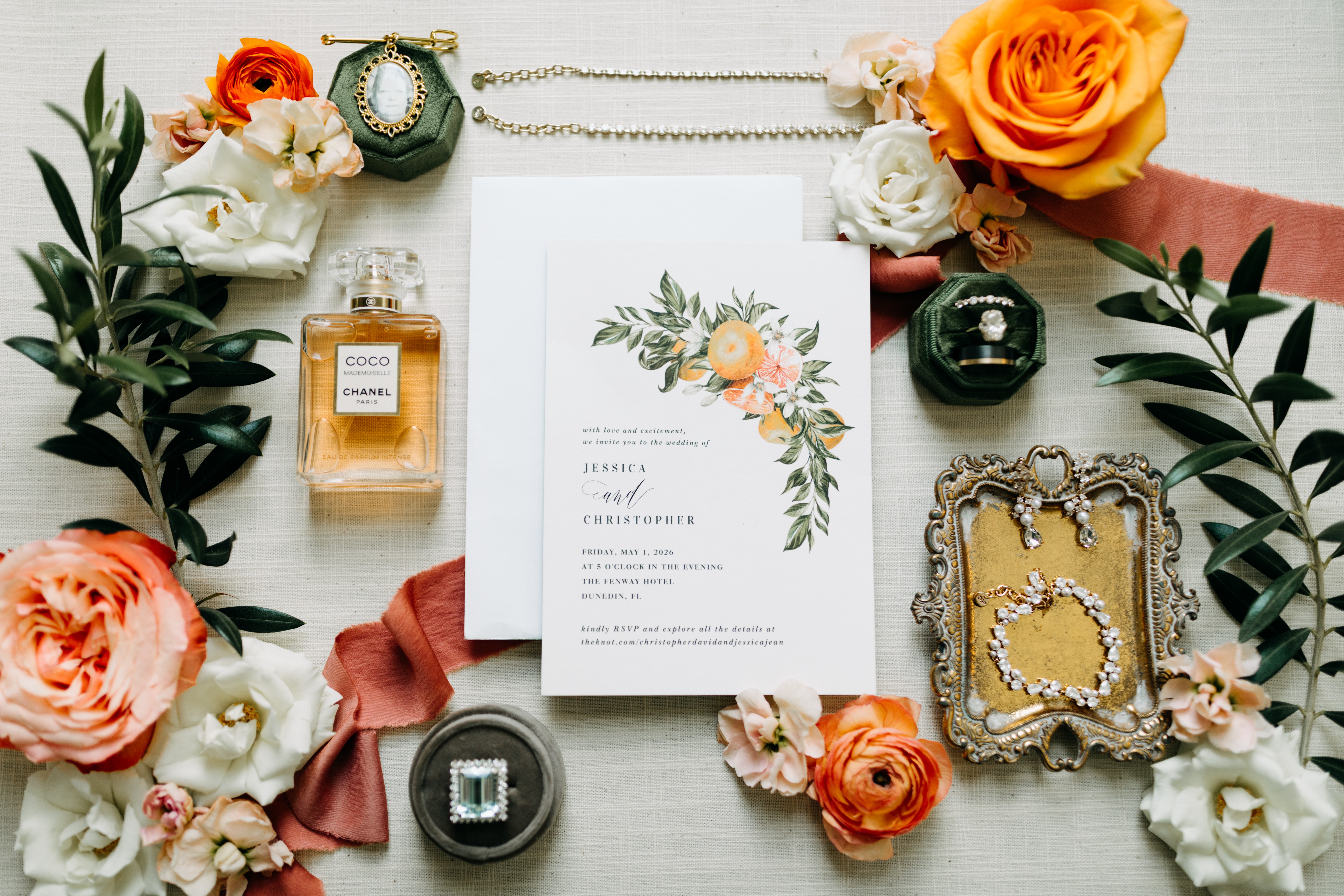 Wedding invitation surrounded by flowers, perfume, jewelry, and decorative items on a white surface.
