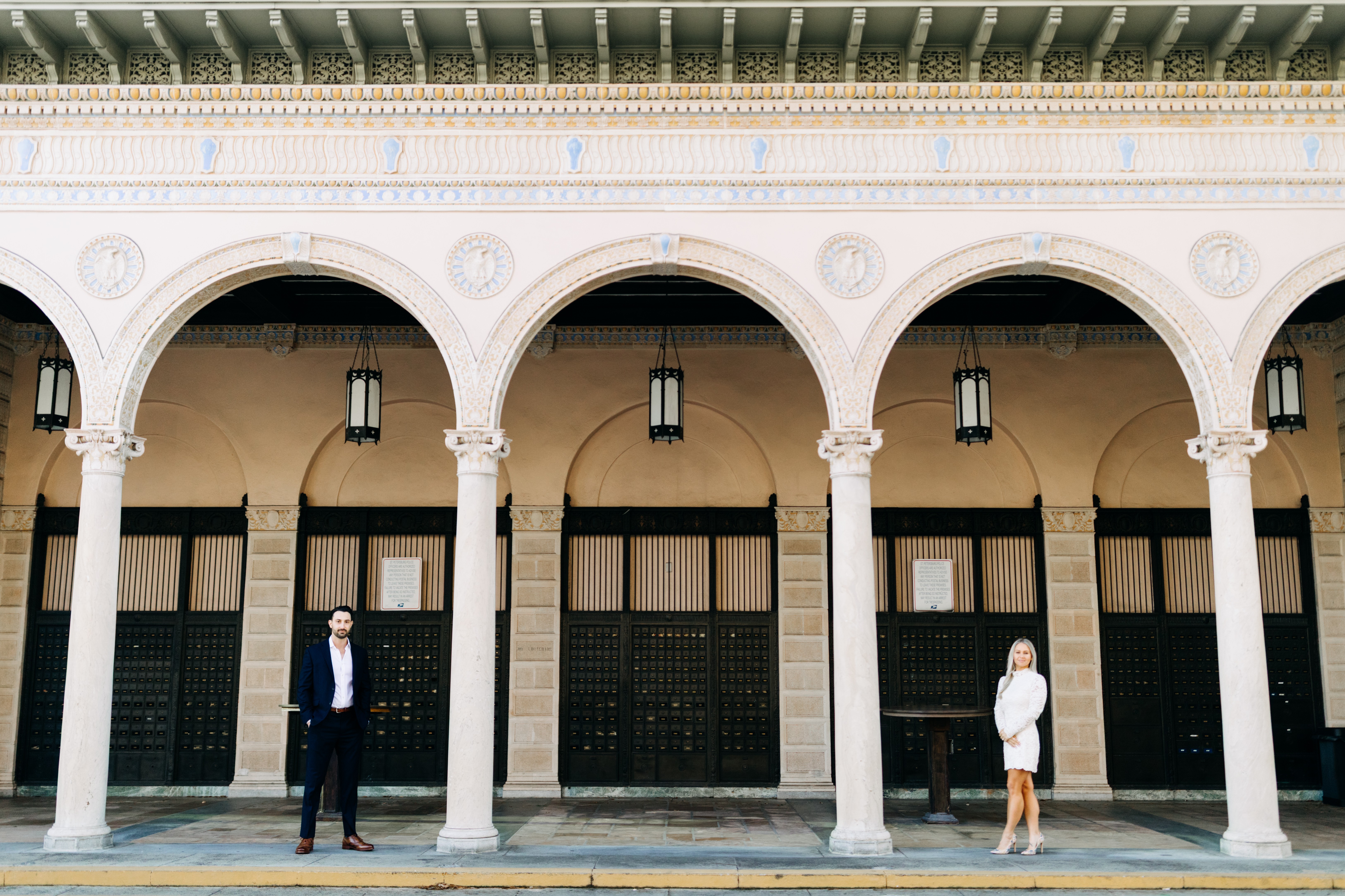 Three arches with columns and hanging lanterns, building facade with closed doors and windows, two people standing in front.