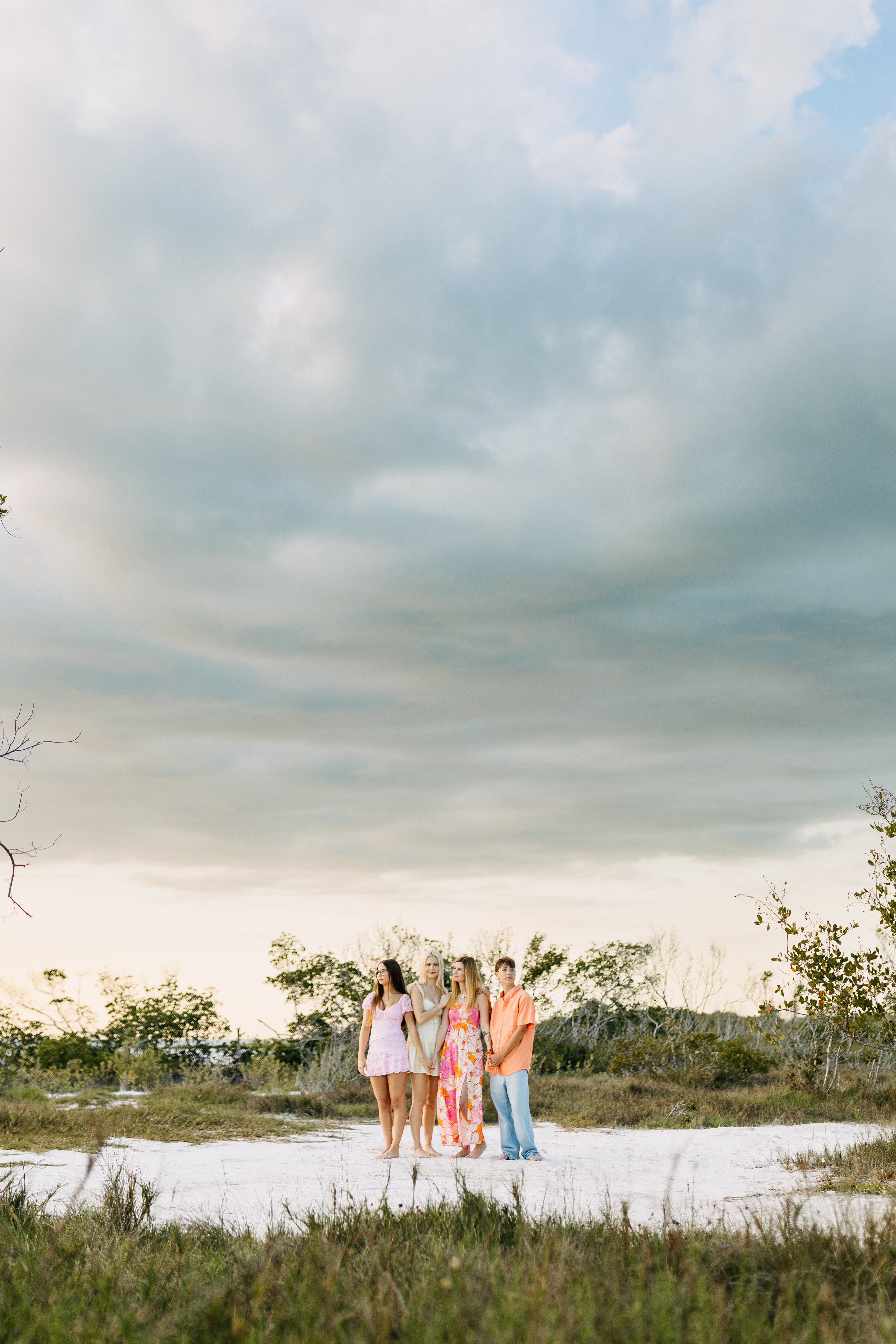 Group of four people standing outdoors on sandy ground with trees and cloudy sky in background.