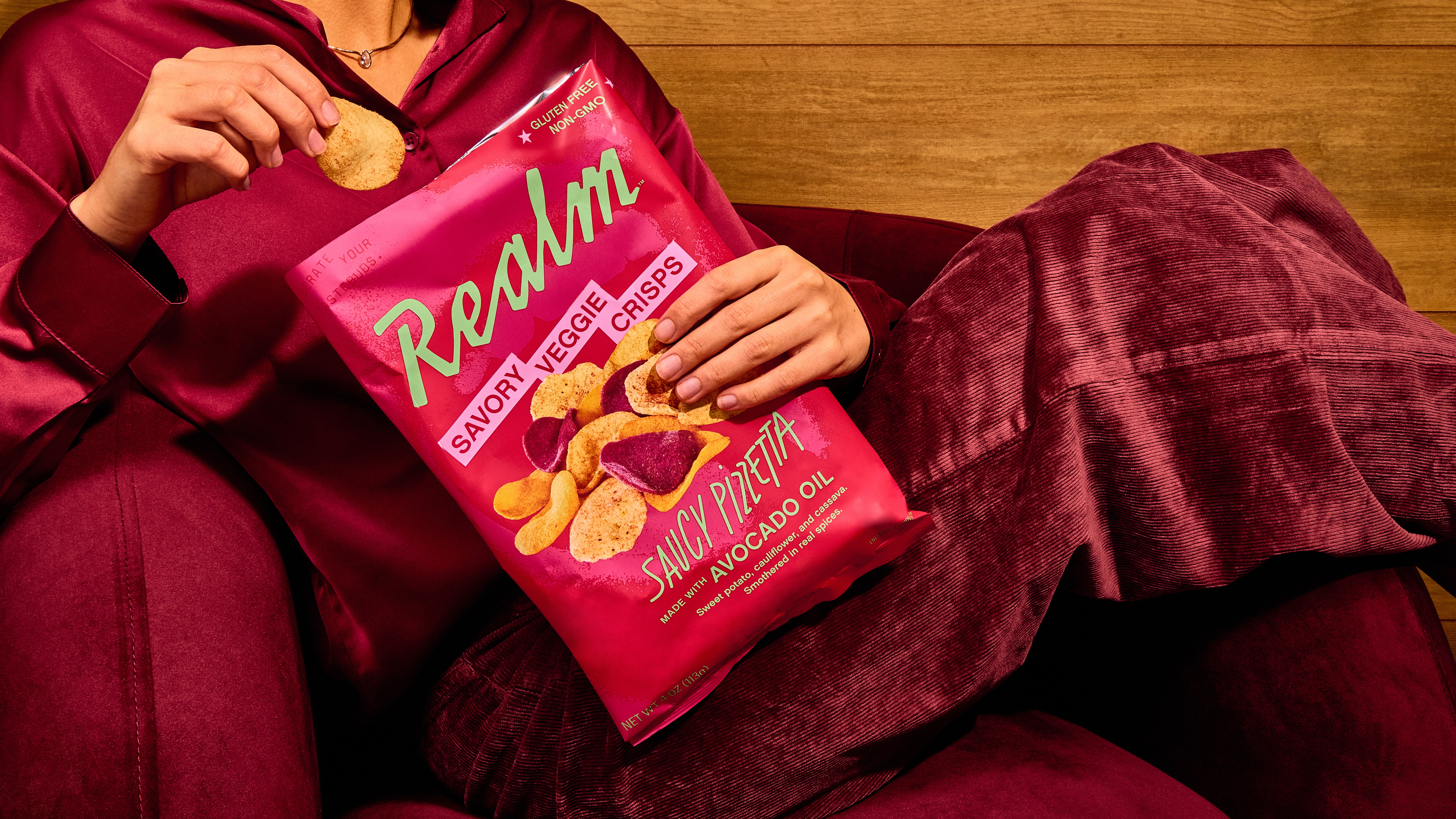 Person in maroon hoodie and pants holding a bag of Ralrm snack chips, sitting on a wooden surface with a blanket.