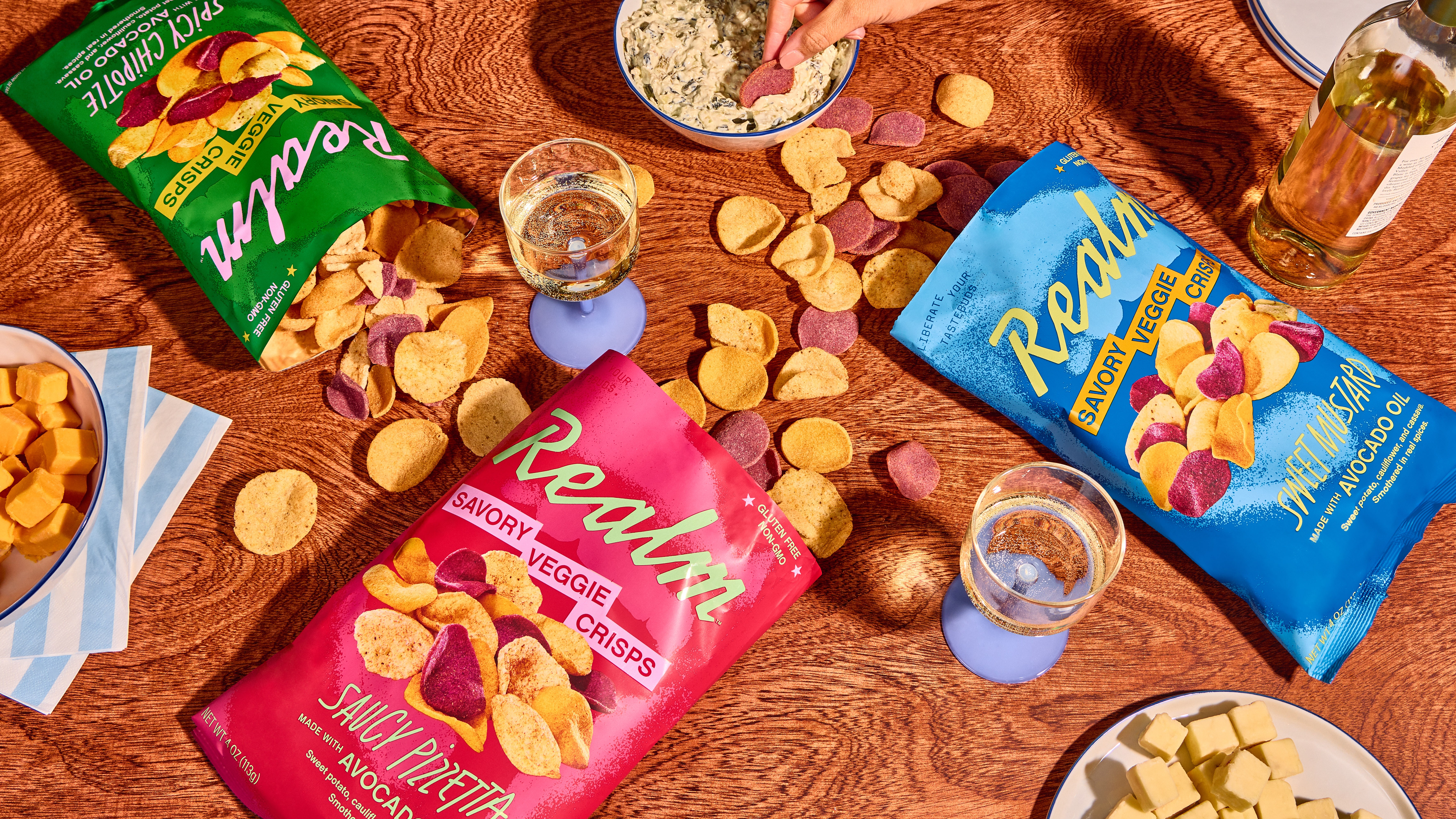 Snacks and chips on a wooden table with glasses of water, including pink and blue bags of Realka chips, and a green bag of Lay's chips.
