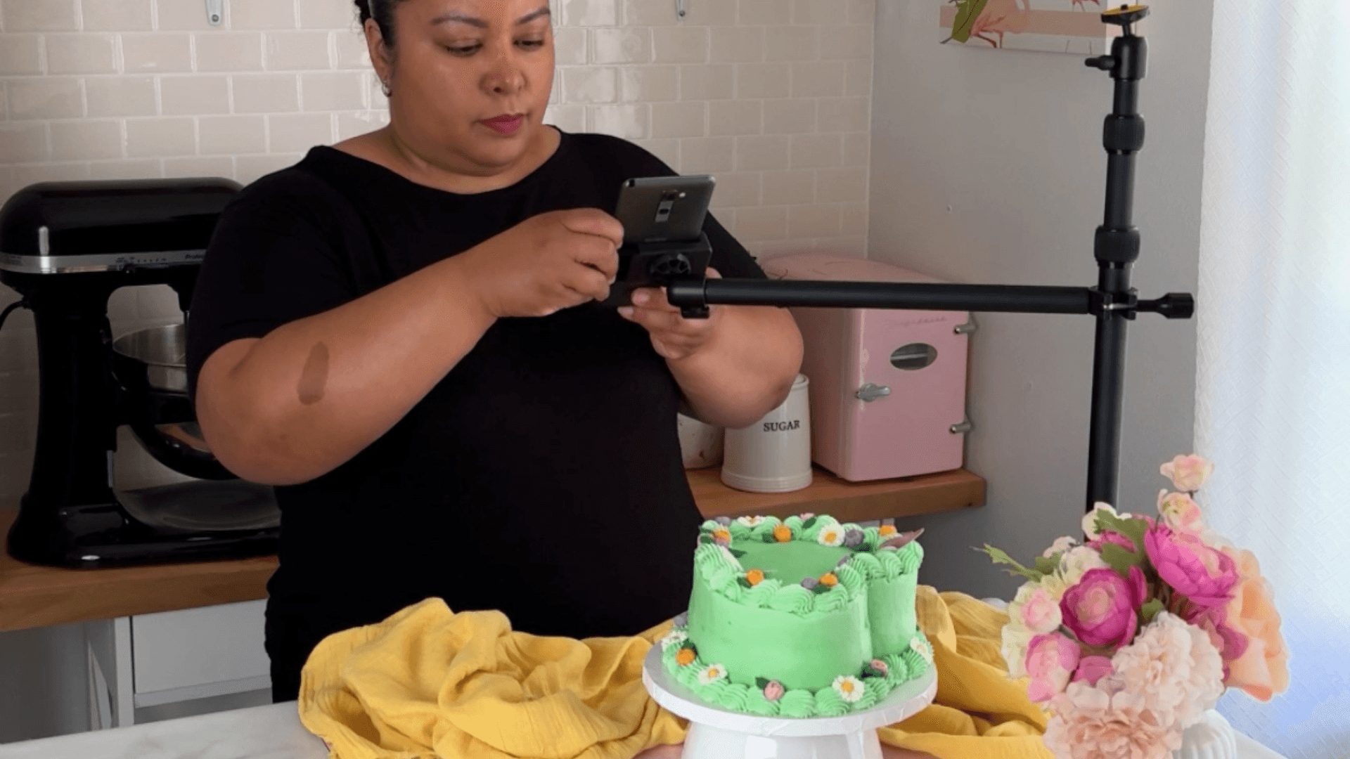 Woman in black shirt taking a photo of a green decorated cake with pink flowers, on a table with yellow cloth, in a room with white brick wall.