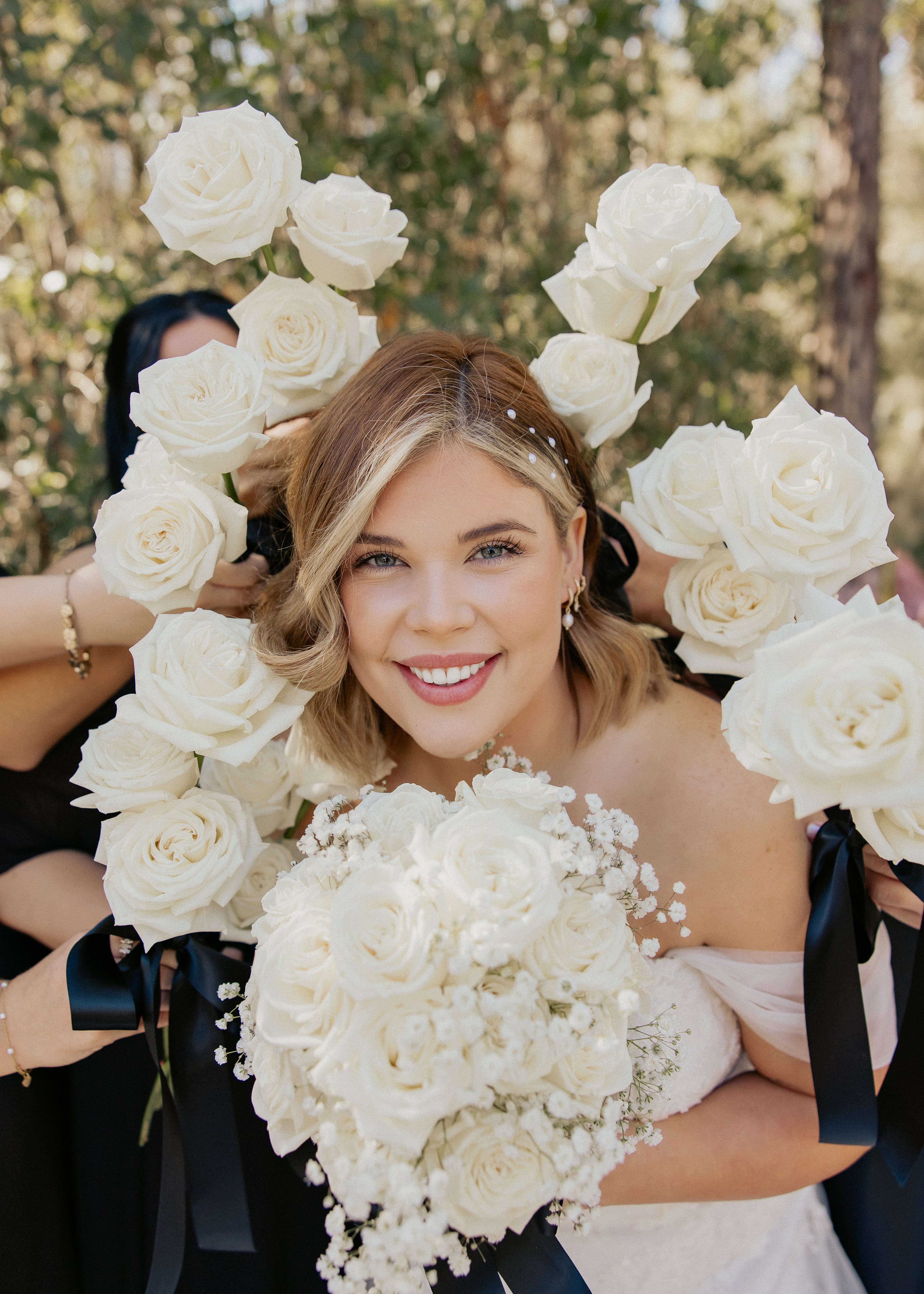 Young woman with short blonde hair smiling, surrounded by white roses and flowers, outdoors with trees in background.