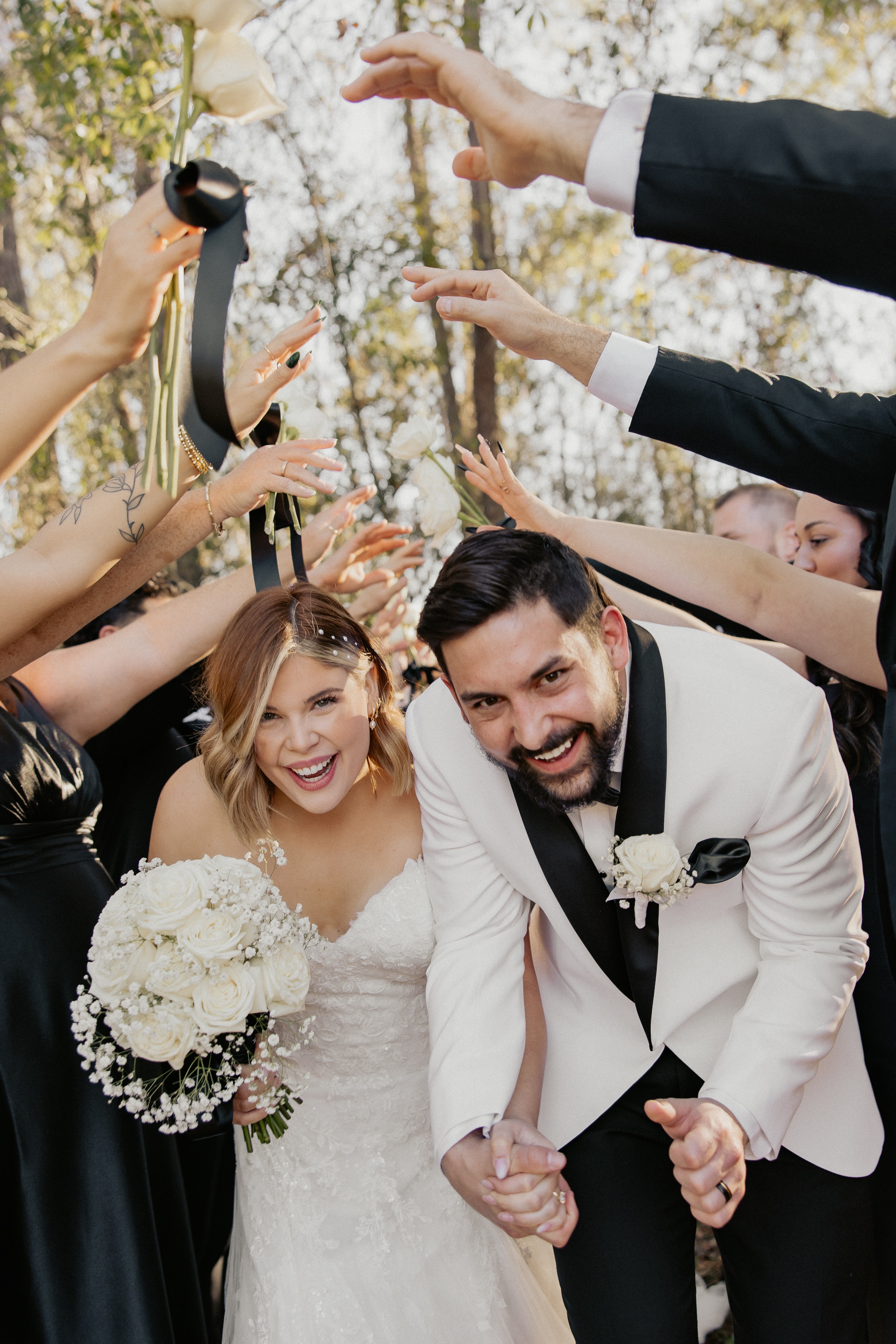 Bride and groom smiling, surrounded by guests raising hands in celebration outdoors, trees in background.