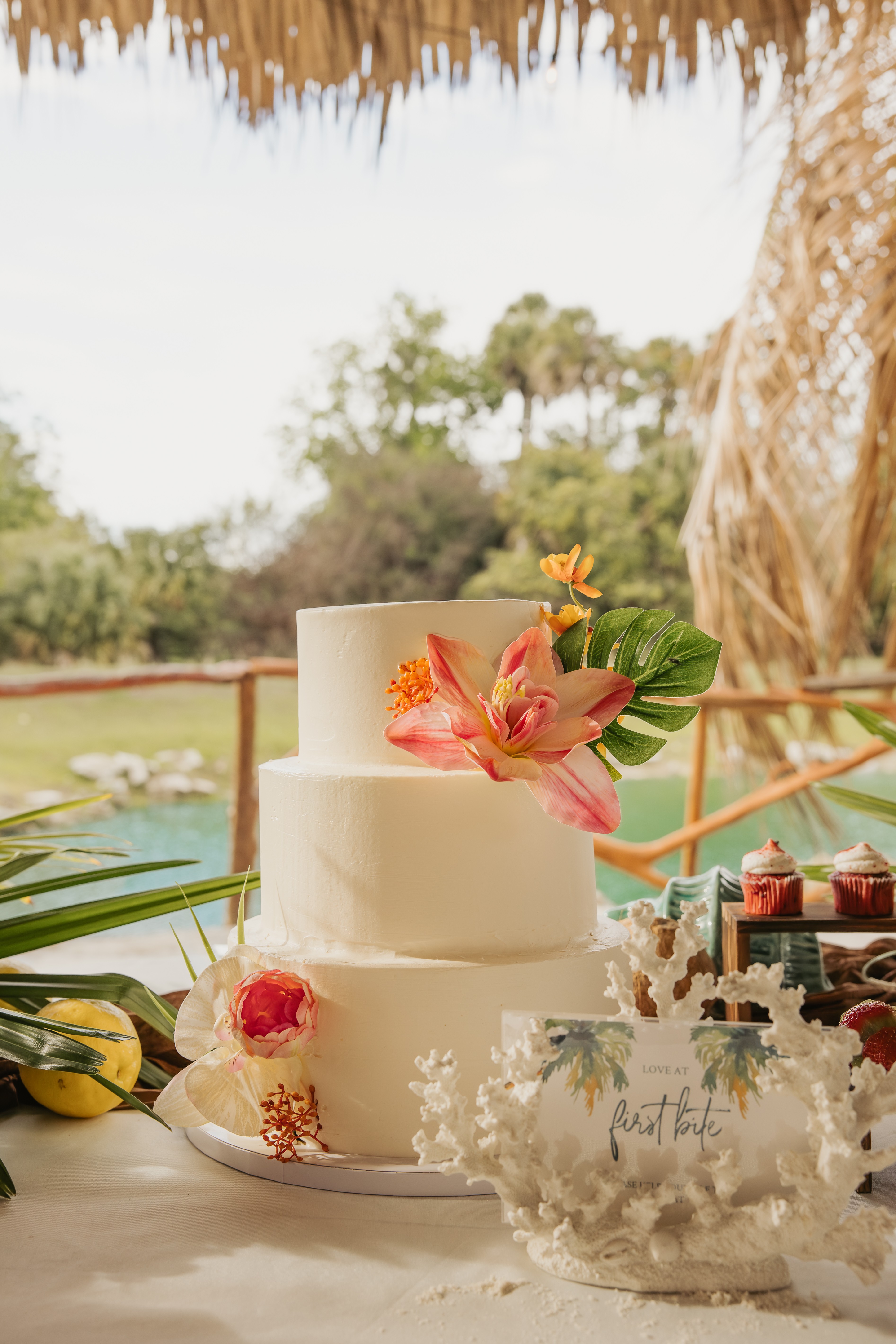 Two-tiered white wedding cake decorated with pink and orange flowers, set outdoors with trees and a thatched roof in background.