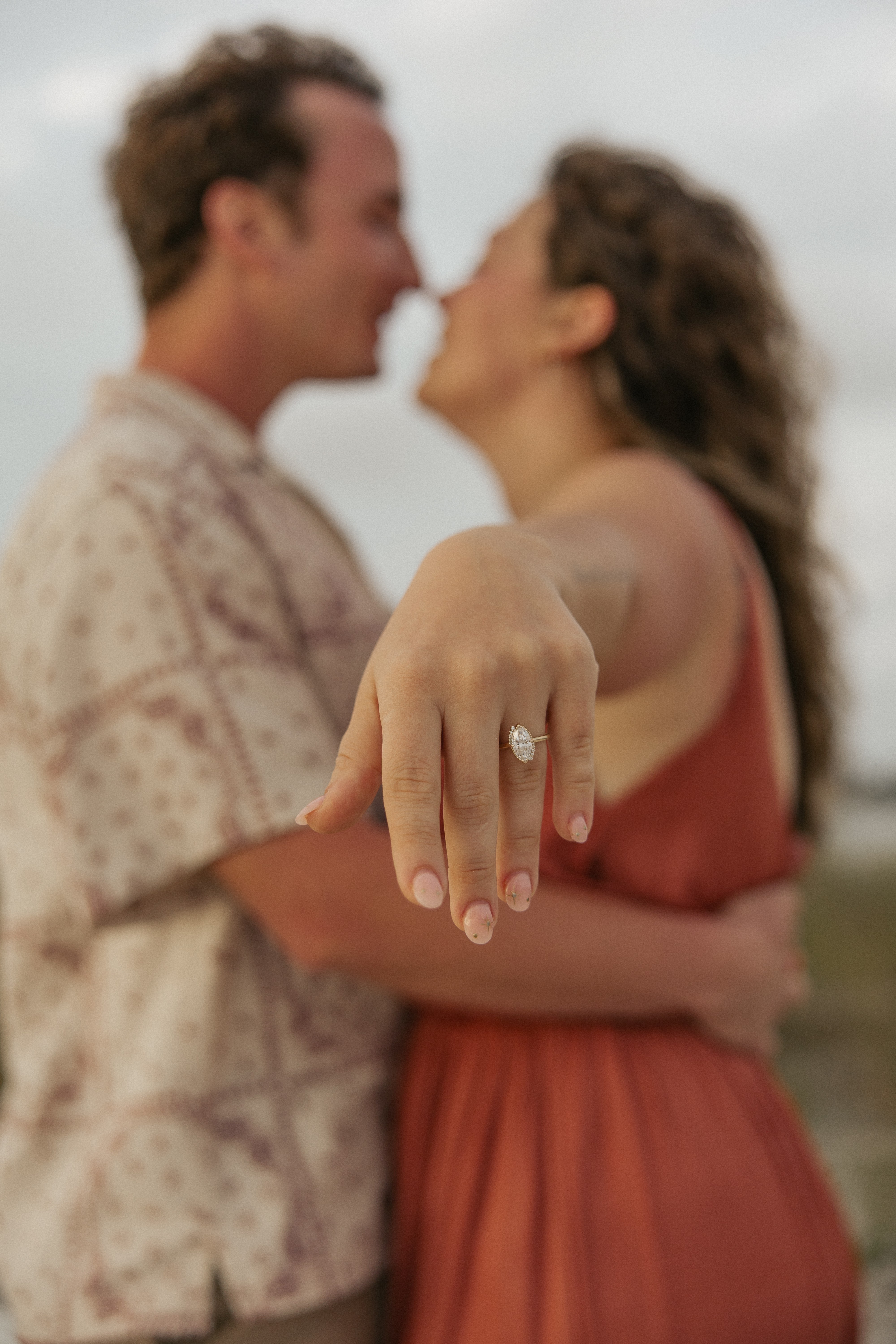 Close-up of a woman's hand with an engagement ring, with a couple kissing in the background.