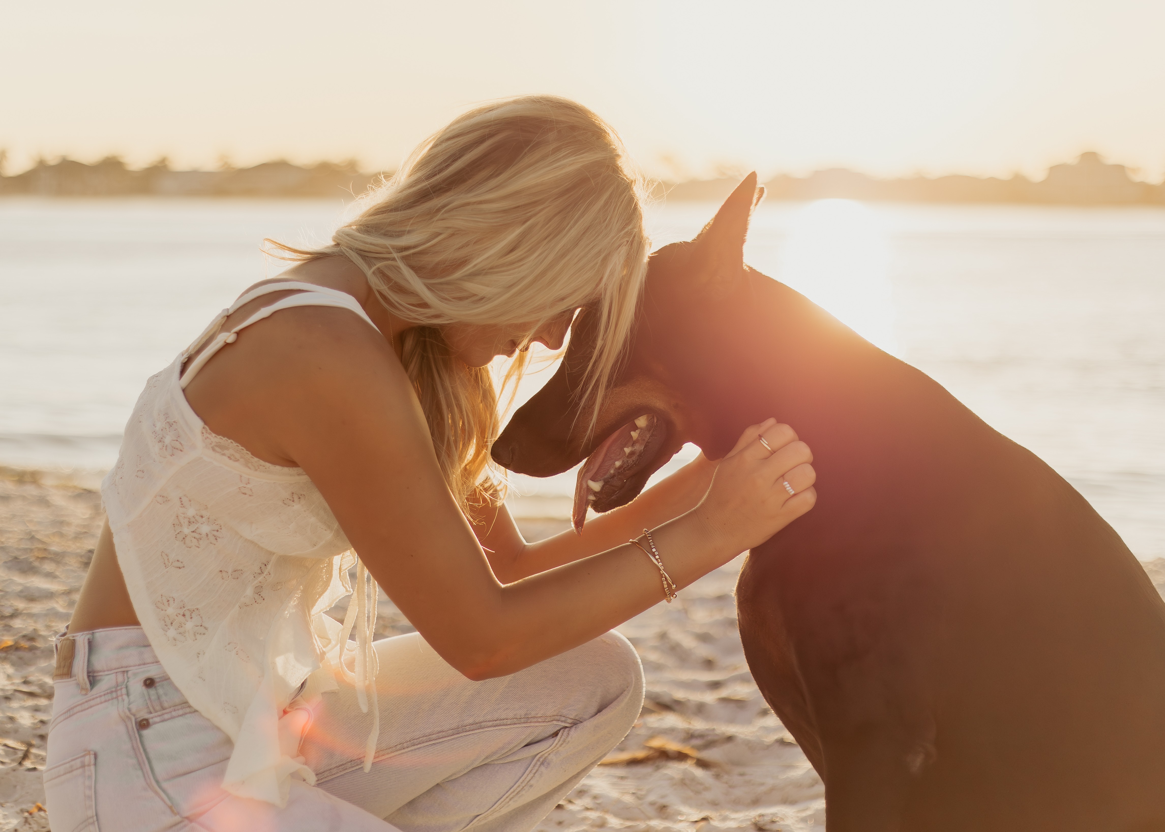Woman with blonde hair gently touching a horse's face on a beach at sunset.