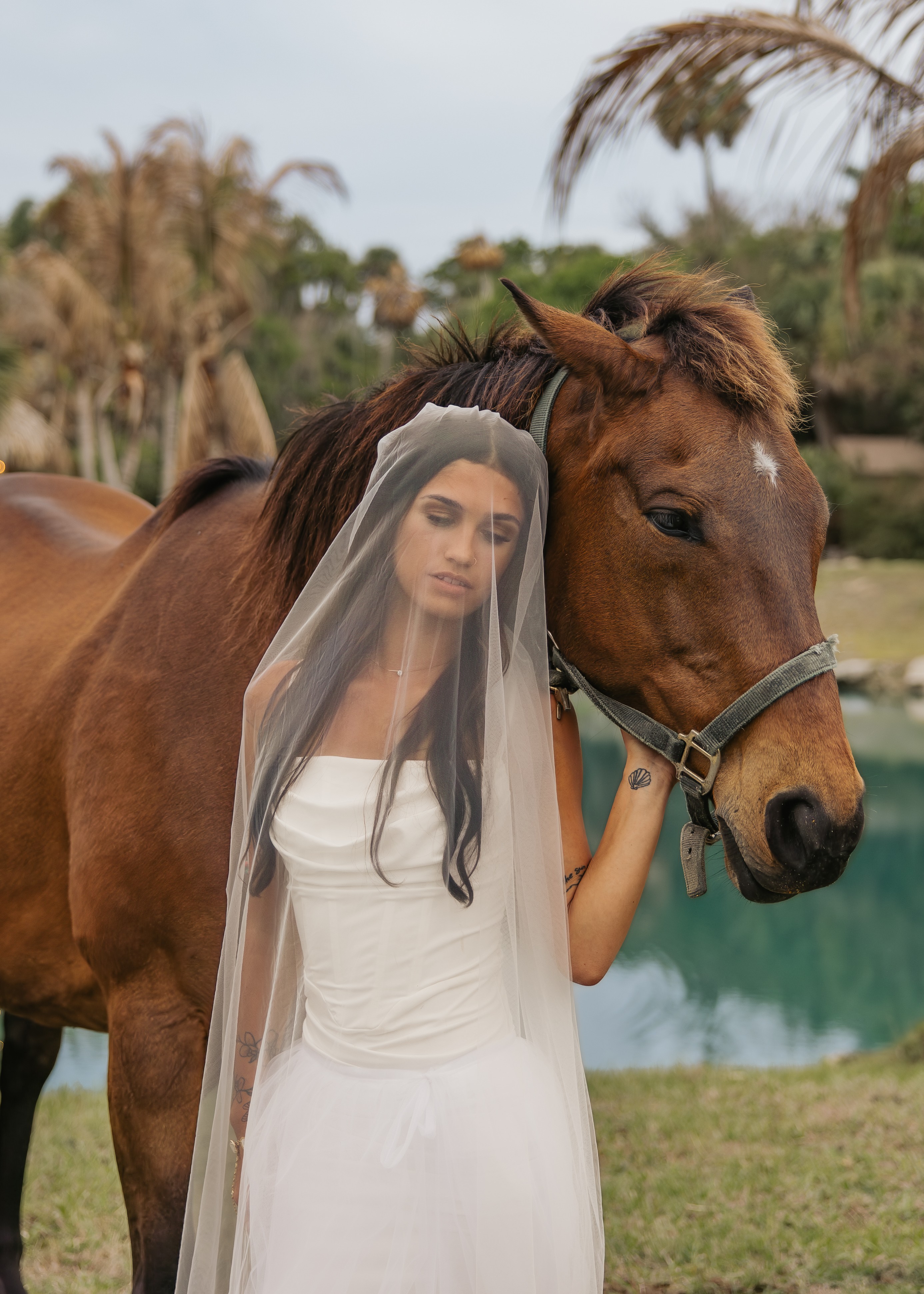 Woman in white dress with veil standing next to a brown horse outdoors near water, trees, and a cloudy sky.