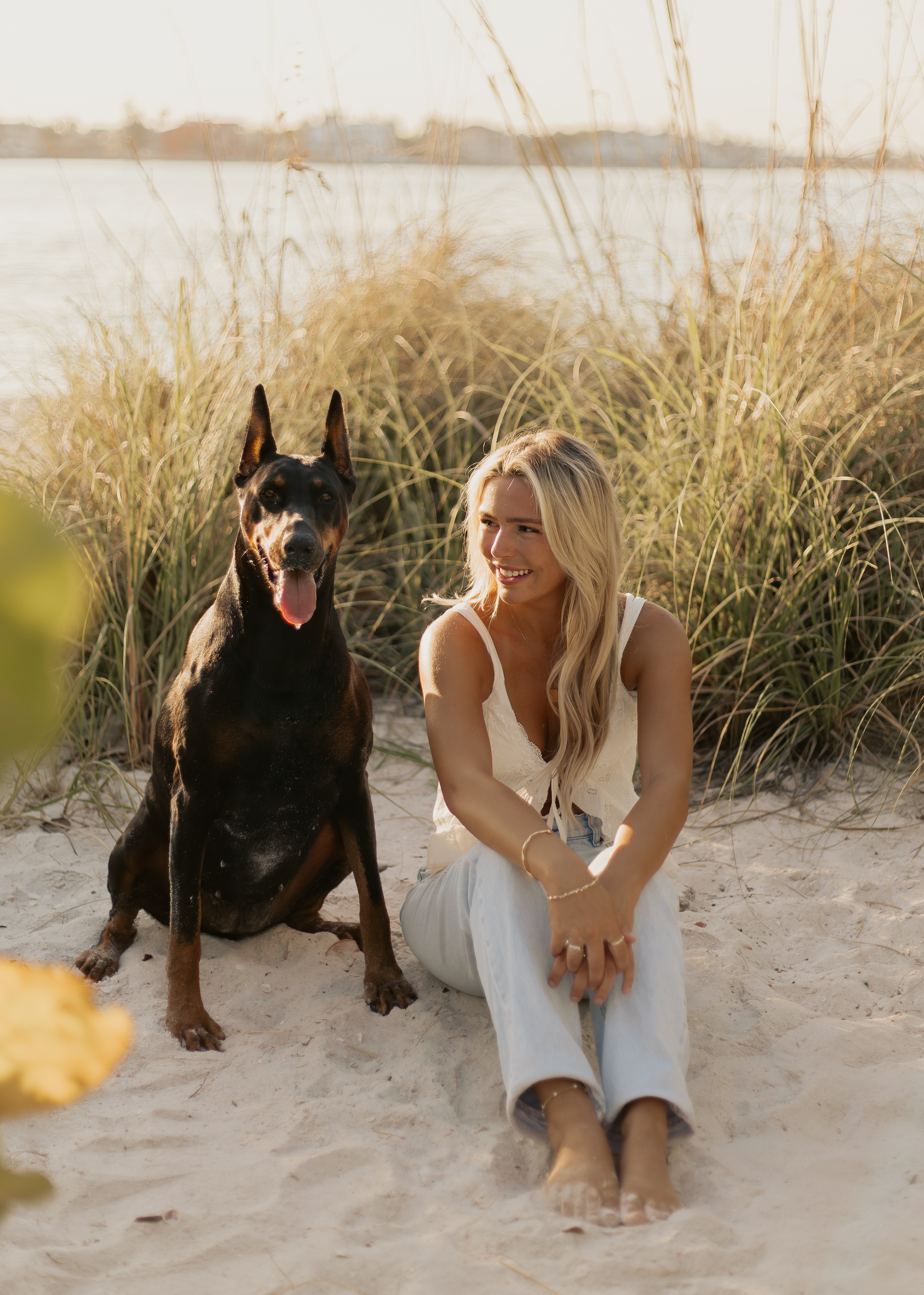 Woman sitting on sandy beach with tall grass behind her, smiling, with a large black dog sitting beside her.