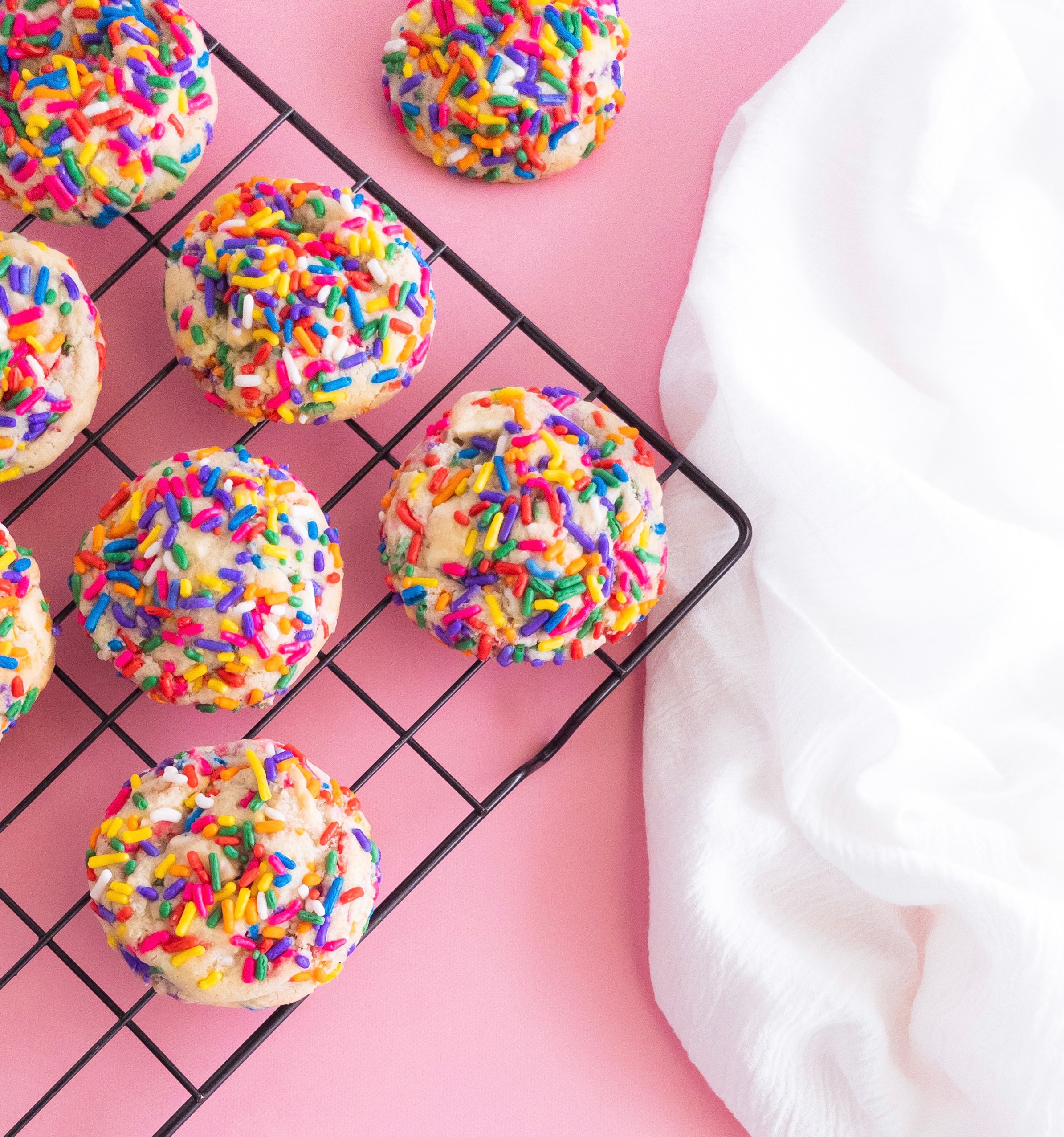Colorful sprinkled cake pops on a cooling rack with a pink background and white cloth, close-up view.