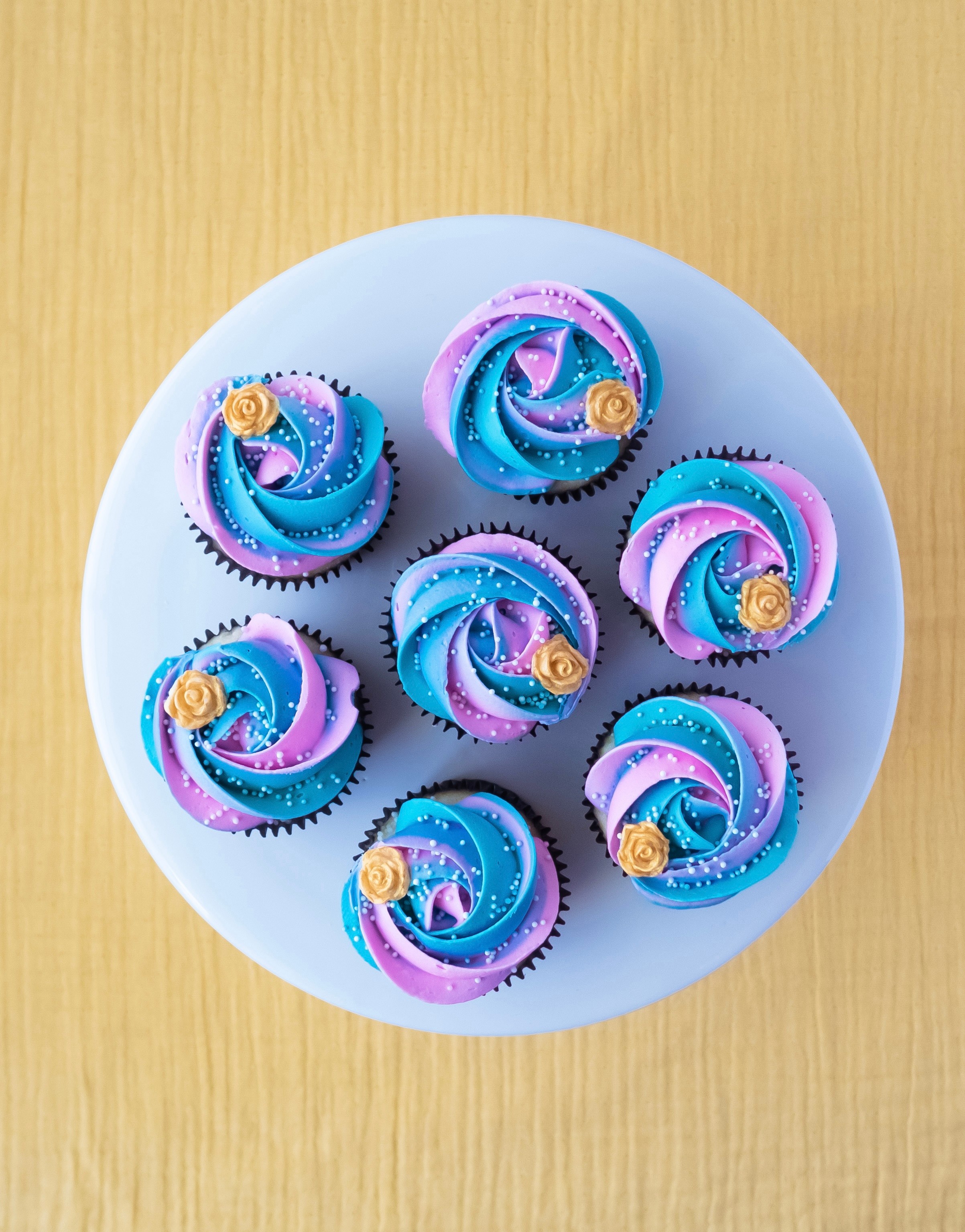 Top-down view of eight cupcakes with blue and purple swirled frosting and small yellow decorations on a white plate.
