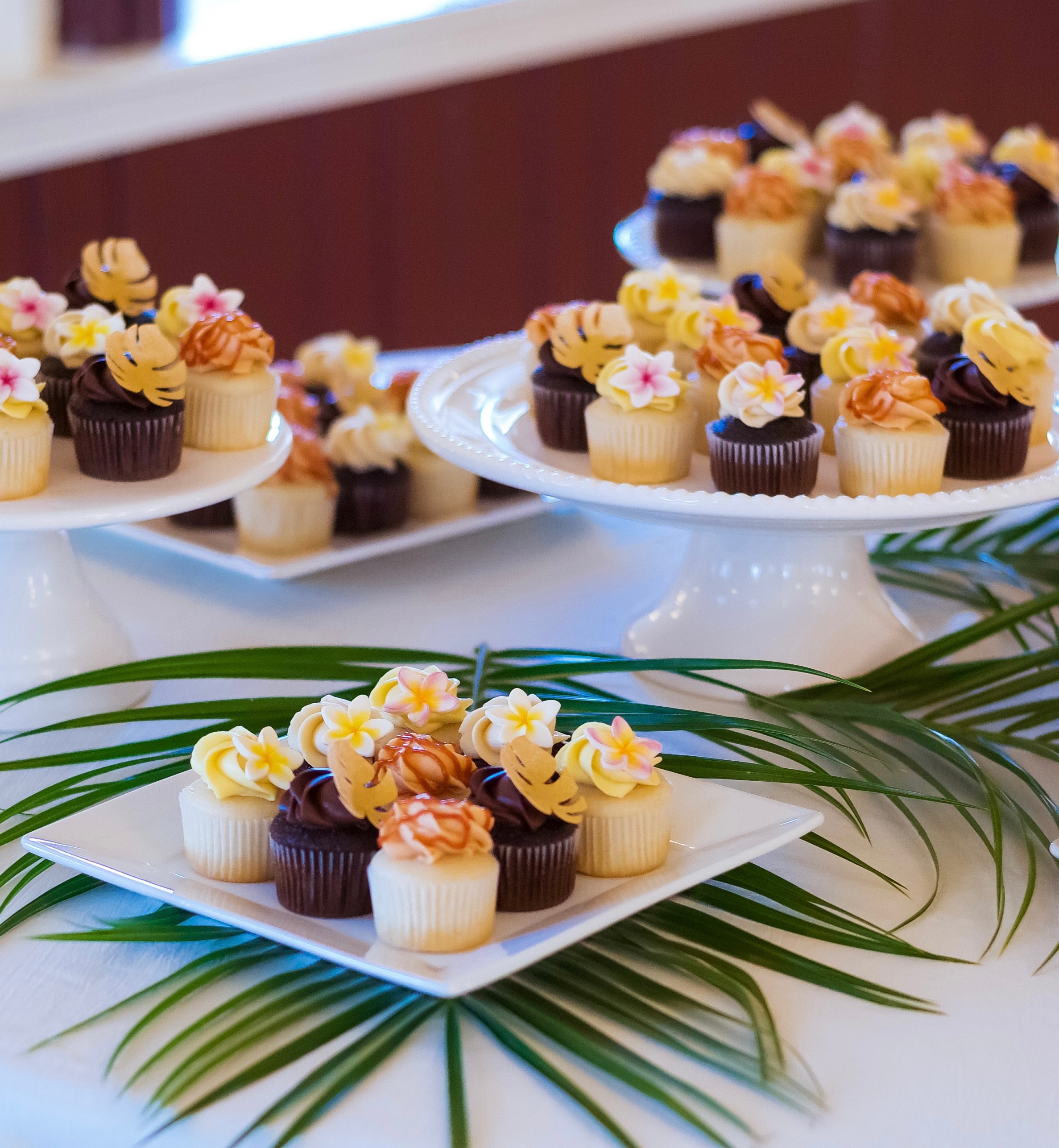 Assorted cupcakes with flowers on white plates, decorated with yellow, pink, and purple frosting, surrounded by green leaves.